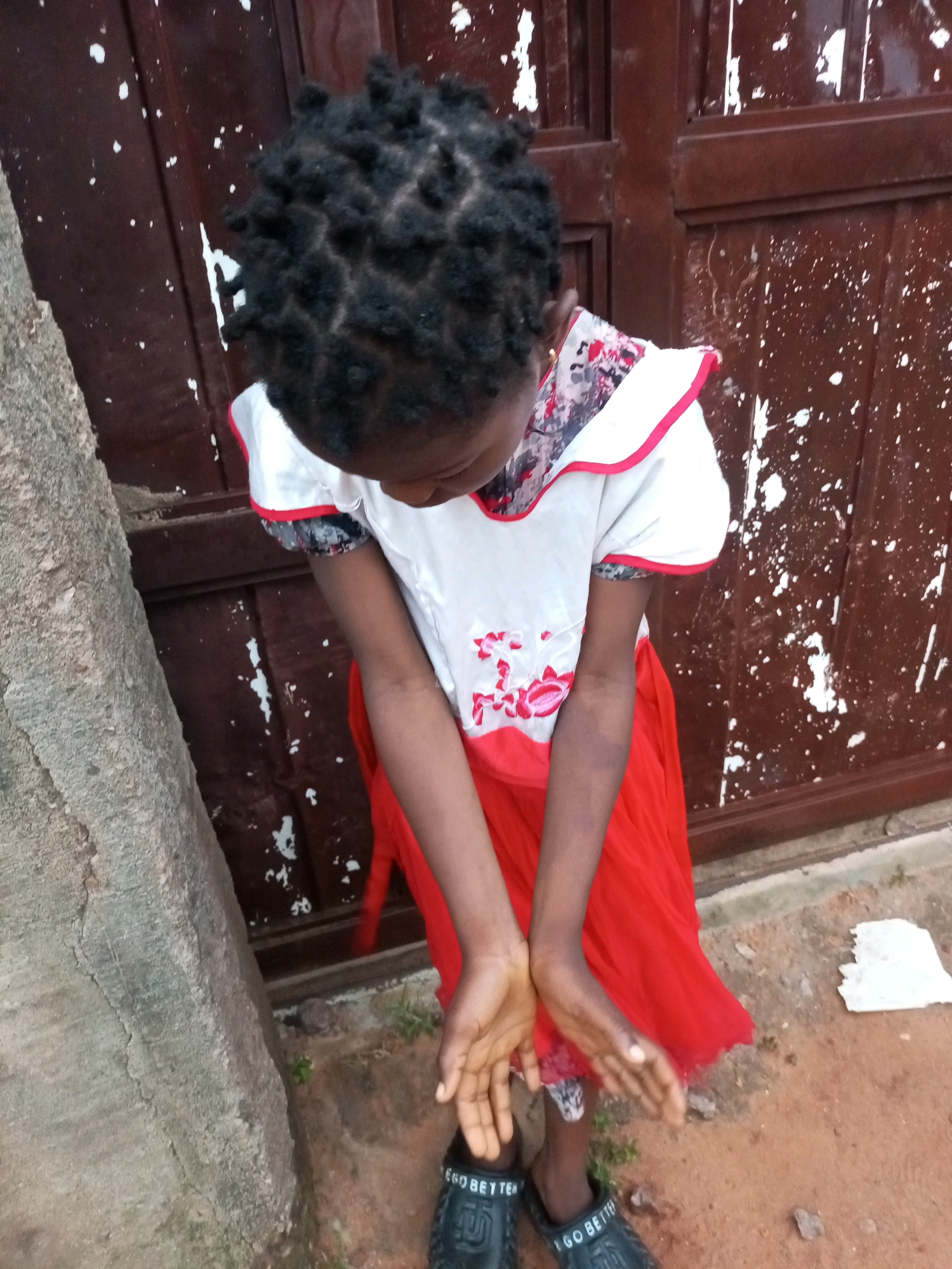 In Tempure, Nigeria, a young girl with dark, braided hair stands outside. She wears a white dress with red trim and floral accents, paired with a flowing red skirt. Her arms are bent at the elbows, with her hands held open and palms up, suggesting she is offering something or waiting to receive it. She is looking downwards. Her feet are clad in black sandals with white lettering that reads "I GO BETTER." The background consists of a weathered, brown door with white splotches and a textured, greyish tree trunk to her left. The ground beneath her is composed of reddish-brown dirt.