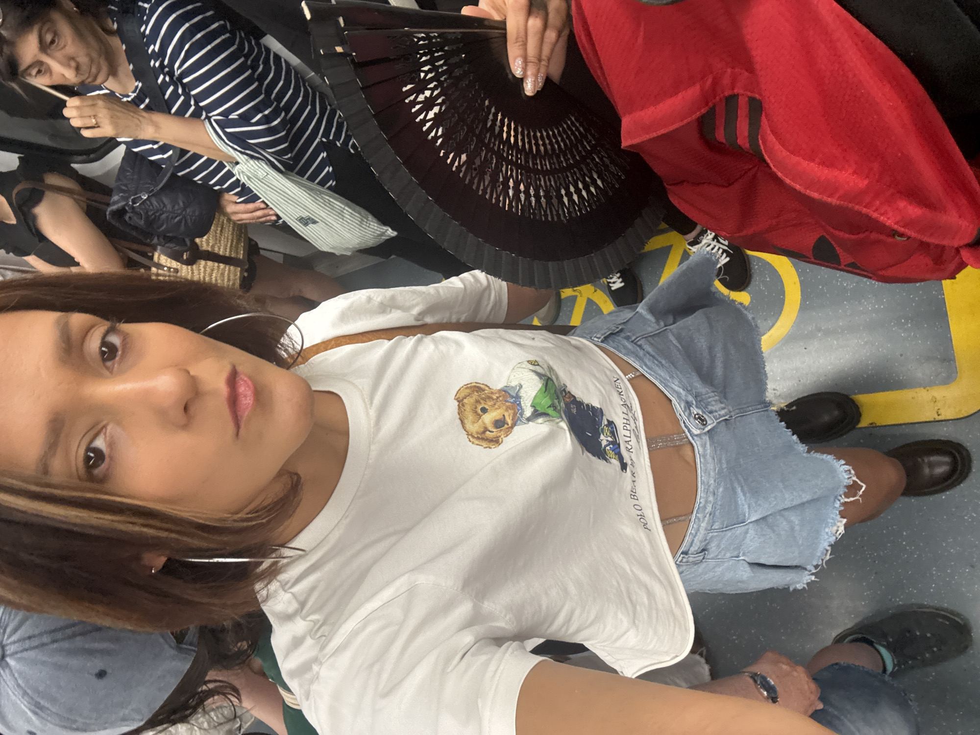 This is a first-person perspective, medium-shot, indoor photograph taken on public transportation, likely a subway or train, in Barcelona, Spain. The main subject is a woman with medium-length brown hair, wearing a white crop top with a Polo Bear by Ralph Lauren graphic and a light-wash denim pleated mini-skirt with a frayed hem. She is also wearing a sparkly chain belt and dark-colored loafers. Her left hand is visible, holding a black folding fan.
In the background, other passengers are visible. To the left, a person wearing a light blue cap is partially visible. Behind the main subject, to the right, an older woman with dark hair and a striped blue and white shirt is seated, holding a ring on her left hand. Another person is wearing a black top and carrying a woven straw bag and a black quilted bag. The floor of the transport is gray with yellow markings. The lighting appears to be artificial, typical of an indoor public space.