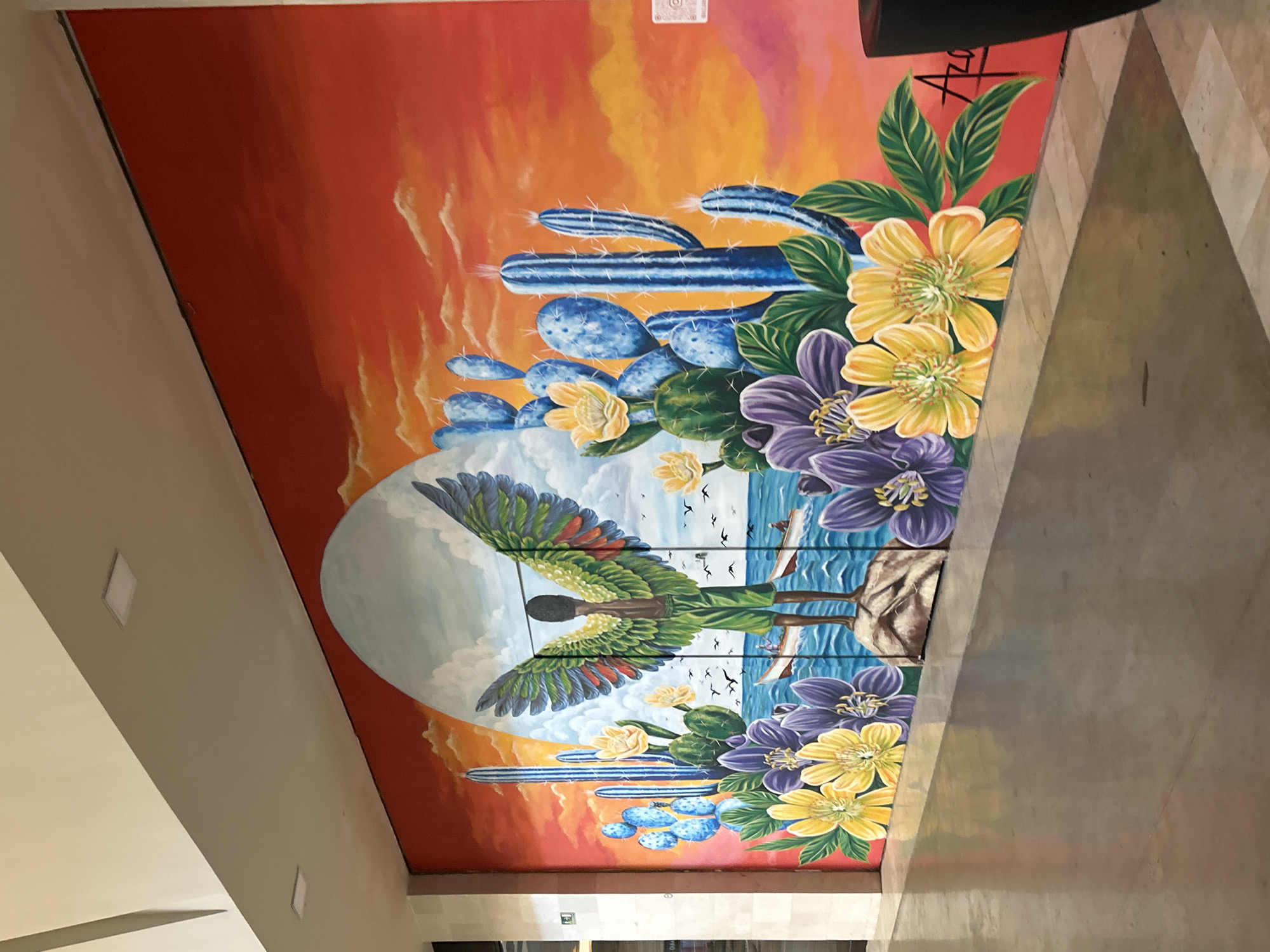 The image shows a vibrant, large-scale mural painted on an interior wall. The mural depicts a surreal desert landscape at sunset, blended with a coastal scene. In the foreground, colorful flowers, including yellow and purple blossoms, are painted, along with tall, blue cacti. A prominent central figure stands on a rocky surface, depicted as a young person with large, green and yellow wings, facing away from the viewer. Behind this figure, a bright sun or moon is set against a gradient orange and red sky with wispy clouds. The sea is depicted in the background with a small boat carrying two figures. Birds are scattered across the sky. To the left of the main scene, there are more cacti and a partially visible person in a doorway. The mural is located in Pampatar, Venezuela. The floor in front of the mural appears to be polished concrete, reflecting the colors of the artwork. A dark trash receptacle is visible to the far right of the image.