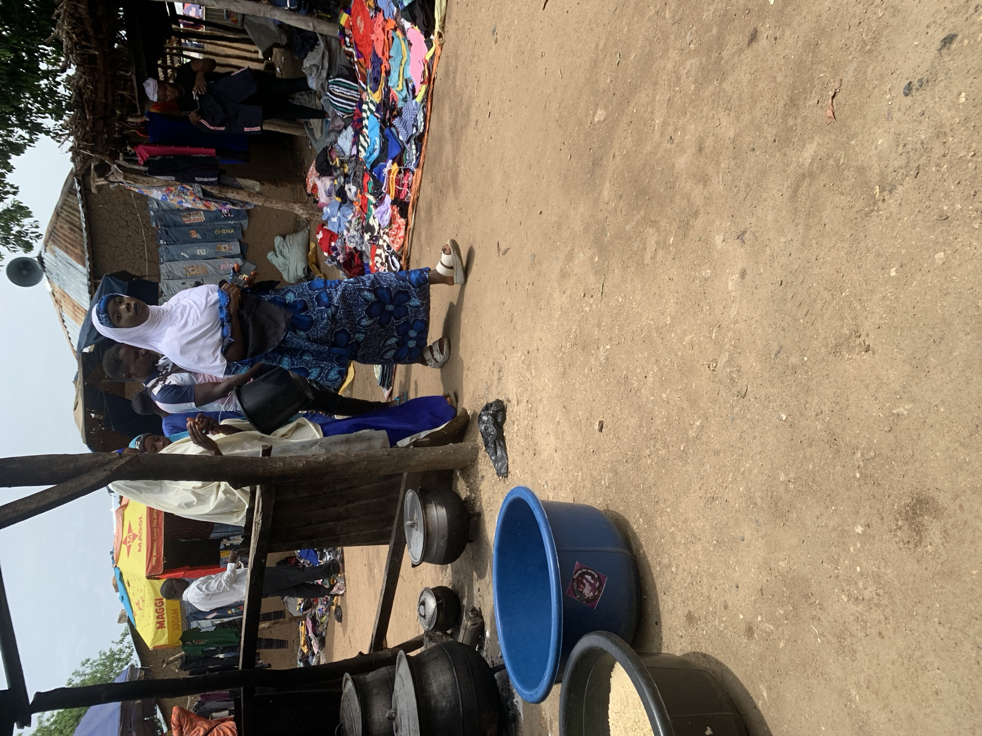 The image depicts a bustling outdoor market scene in Nigeria, likely during the daytime with the sun casting shadows. In the foreground, a woman wearing a white hijab and a blue patterned dress stands in a dirt-floored market. She is looking towards the viewer and holding a small red object, possibly fruit, in her hand. Beside her, several large metal cooking pots and a large blue plastic basin are visible, suggesting food preparation or sale.
In the background, a stall is laden with a colorful assortment of clothing, with items neatly folded or hung. More clothing is visible in piles to the left of the stall, creating a vibrant and busy atmosphere. Several other people are present in the market, including individuals appearing to be vendors or shoppers interacting with the goods. One person in the background is wearing a white shirt and dark trousers, seemingly engaged in an activity related to the stall. A yellow awning with "MAGGI" branding is prominently displayed on one of the stalls, indicating a food product brand. The overall scene conveys a sense of lively commerce and daily life in a Nigerian market.