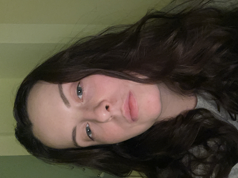 A young woman with fair skin, light eyes, and dark, wavy hair fills the frame. Her face is slightly tilted, offering a direct gaze. Soft lighting highlights her features. A simple grey shirt is partially visible. The background is a muted greenish hue, adding a subtle backdrop to the portrait.