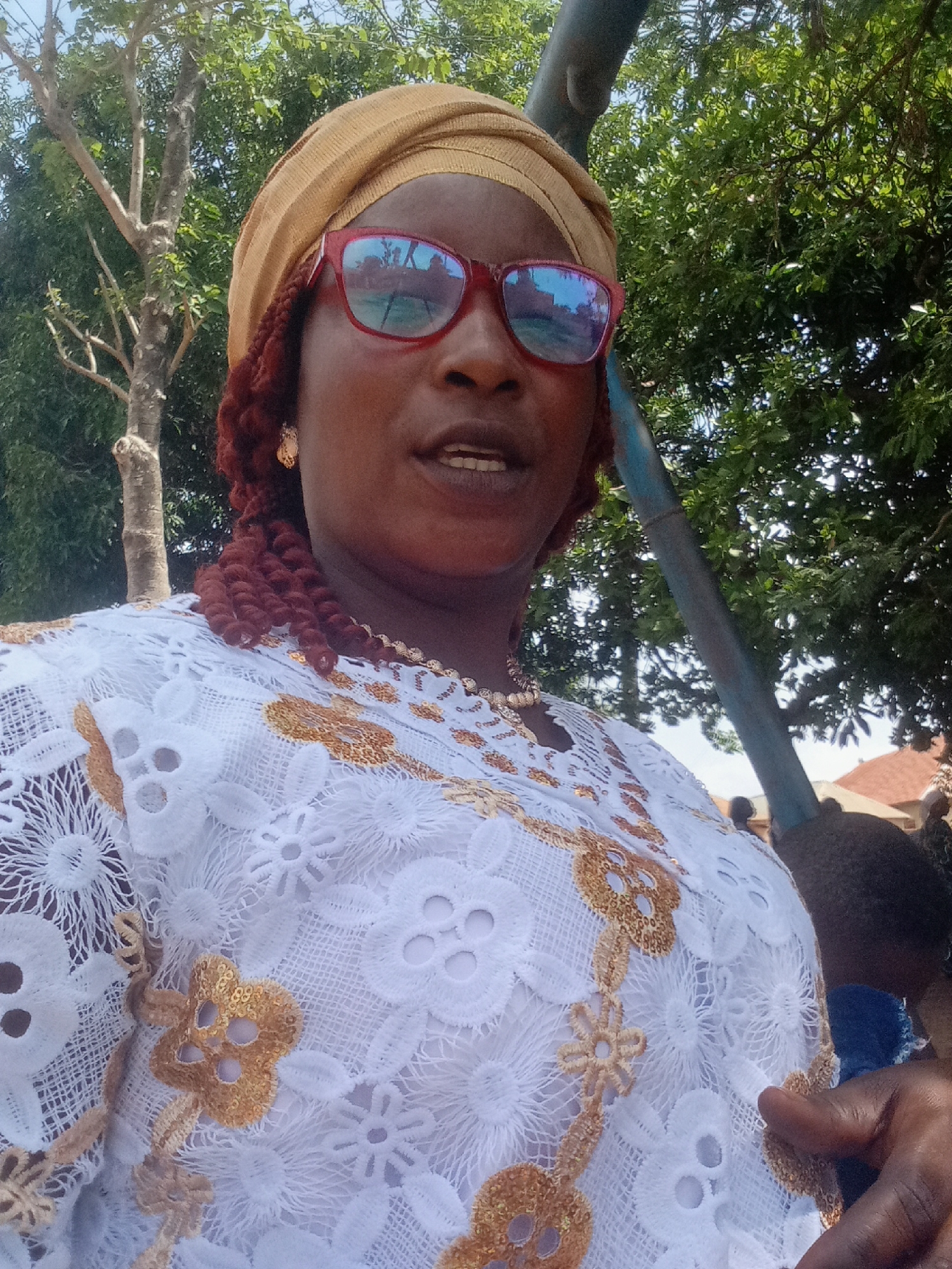 A medium shot captures a person outdoors in Akwanga, Nigeria. The individual wears a light tan headwrap, red-framed glasses with a sky reflection in the lenses, and a white lace blouse adorned with gold floral embroidery. The person has dark skin and reddish-brown braided hair visible near the face. They are wearing gold earrings and a gold necklace. Behind them are trees, a blue pole, and a partial view of a building, indicating an urban or suburban setting. The lighting suggests it is daytime, and the presence of trees hints at a park-like area or residential neighborhood.