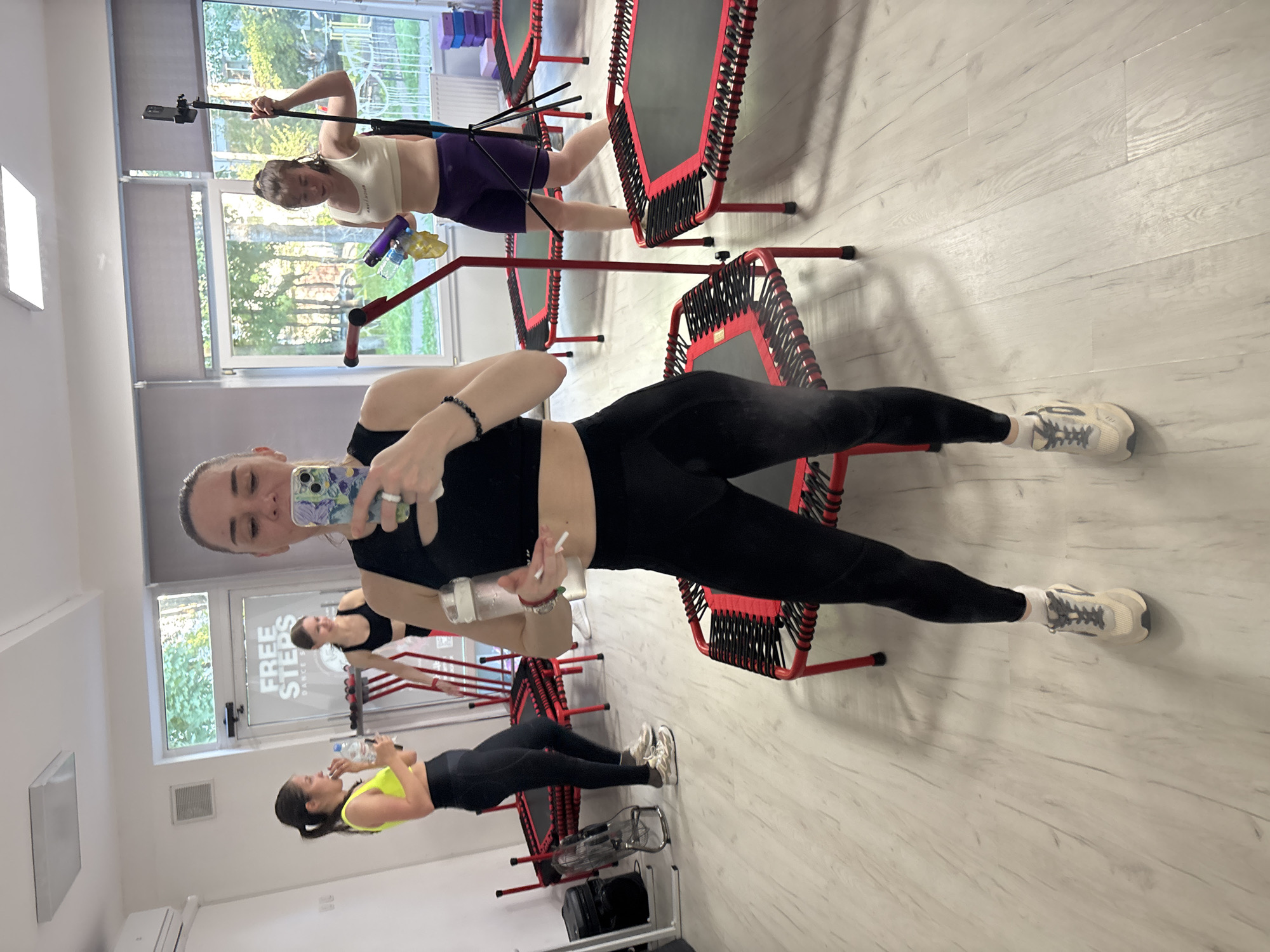 In a brightly lit indoor space, likely a fitness studio in Sinyavino, Russia, three women are engaged in a fitness activity. The room features a light-colored wooden floor and white walls, with large windows allowing natural light to filter in. Several mini-trampolines with red frames and black mats are arranged in the space, suggesting a trampoline fitness class.
In the foreground, a woman is taking a selfie, holding a smartphone in her right hand and a water bottle in her left. She is wearing a black sports bra and black leggings, with light-colored athletic shoes. Her hair is pulled back in a bun.
To her left, another woman, wearing a bright yellow sports bra and black leggings, is holding a water bottle and looking away from the camera. She is also wearing athletic shoes.
Further back, a third woman is visible, wearing a white crop top and dark purple athletic shorts. She is holding several water bottles and appears to be in motion. She is standing near a tripod with a phone attached, possibly recording the class.
In the background, a doorway with frosted glass reveals the text "FREE STEPS DANCE STUDIO" and the city name "Sinyavino." A fan is positioned on the left side of the image, and a stack of purple yoga blocks is visible near the back wall. The overall scene depicts a women's fitness session in progress.
