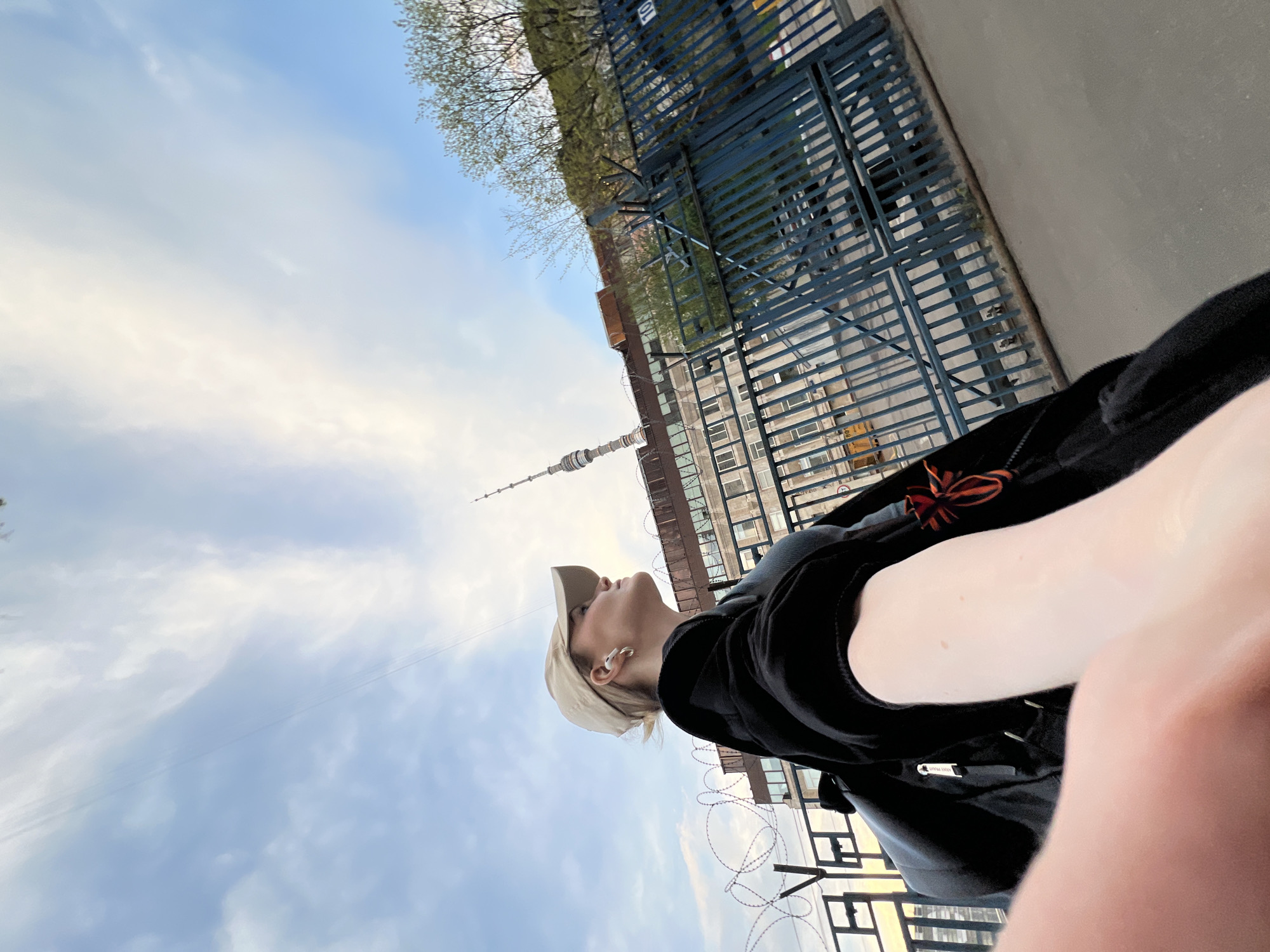 A low-angle selfie shows a person in Khimki, Russia, looking toward the Ostankino Tower in the distance. The person is wearing a light-colored cap, dark clothing and has white wireless earbuds in their ear. A gate and barbed wire are visible in the middle ground, with buildings in the background. The sky is cloudy.