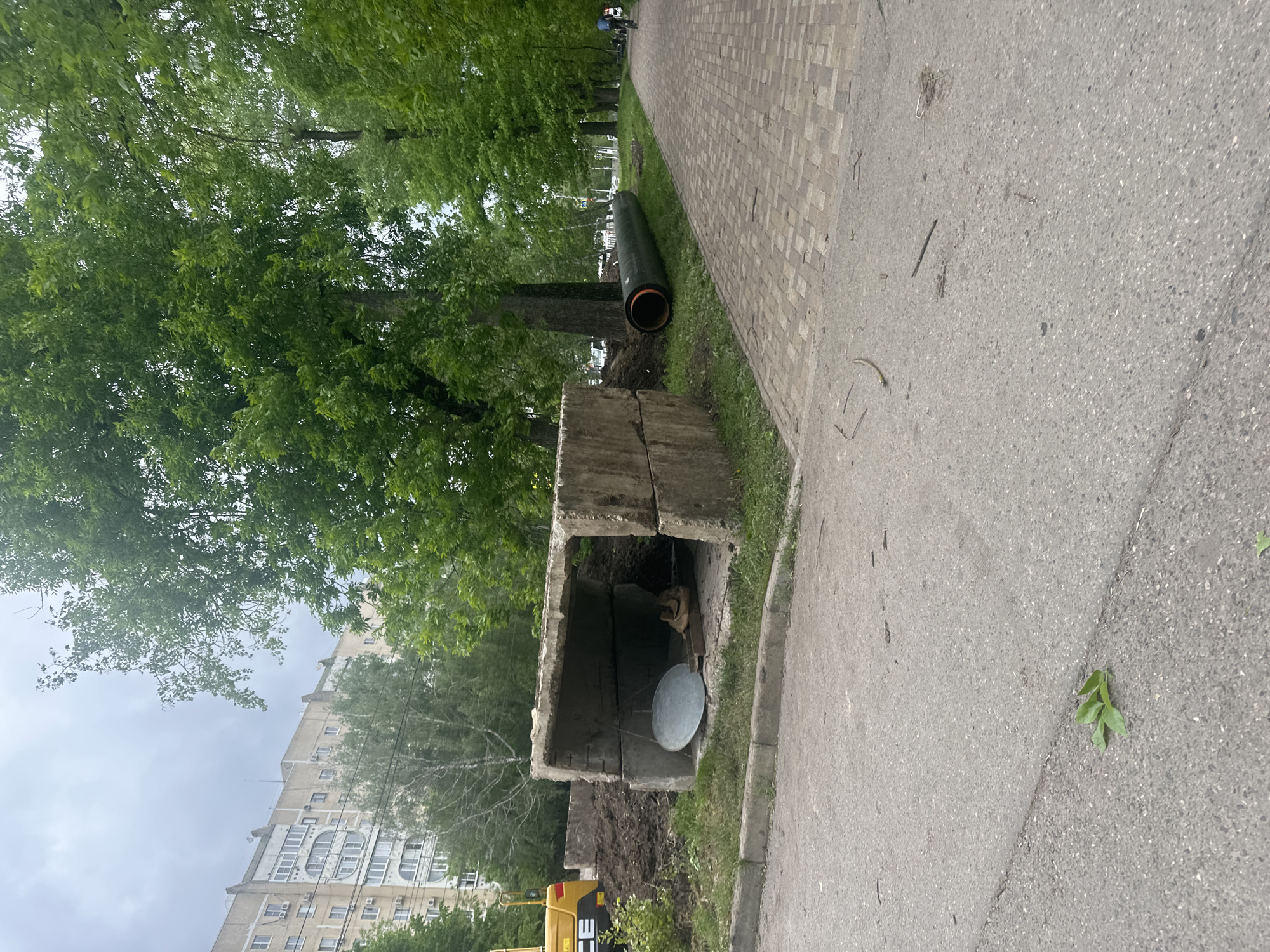 The image depicts a street scene in what appears to be an urban area. A large concrete structure with an open top is situated on the edge of the street, next to a patch of grass. Inside the structure is a metal disc. A large black pipe lies on the grass behind the concrete structure. A sidewalk is visible next to the grass, made of gray rectangular paving stones. A gray asphalt road stretches along the side of the image. Trees and a multi-story building are visible in the background, suggesting an urban setting. A piece of green leaves is on the road. Based on the setting and appearance, the location is possibly Stavropol, Russia.