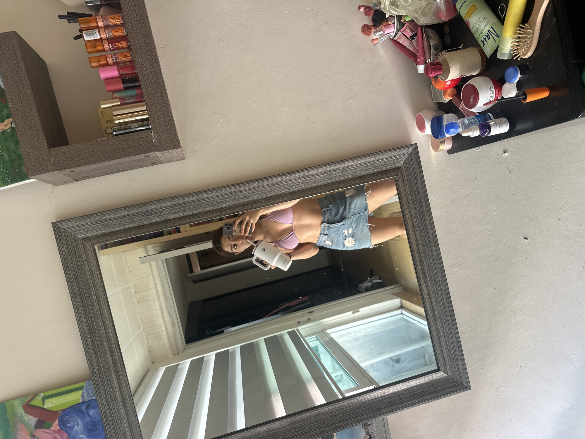 The image shows a medium-sized, rectangular mirror with a grey frame hanging on a light-colored wall. The reflection in the mirror reveals a young woman taking a selfie. She is wearing a pink bikini top and denim shorts. She is holding a white water bottle in her left hand and a phone in her right hand. Behind her, a window with horizontal blinds is visible, showing natural light. Above the mirror, on the wall, there is a shelf filled with various lip glosses and lipsticks. To the right of the mirror, a surface is cluttered with makeup brushes and cosmetic products. The location cannot be determined from this image.