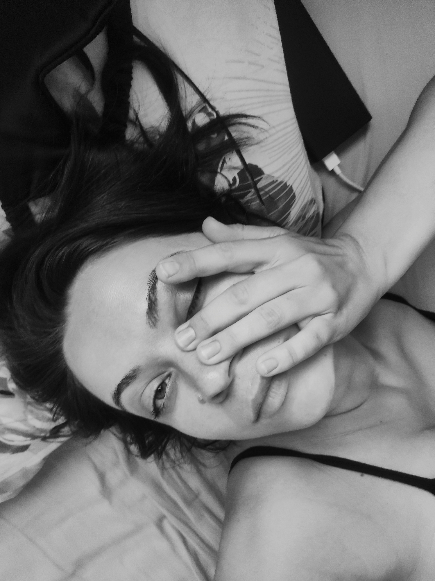 A black and white, high-angle, close-up shot shows a woman lying in bed, with her hand covering her face. Her dark hair is spread across a pillow. The woman is wearing a thin-strapped garment, possibly a camisole or bralette. The pillow behind her has a patterned design. A black object, possibly a tablet or laptop, is visible to her right, with a white charging cable plugged into it. The bedding appears to be light-colored. The scene is indoors, suggesting a bedroom. No specific location in Chaikovsky, Russia, can be identified from the provided image.