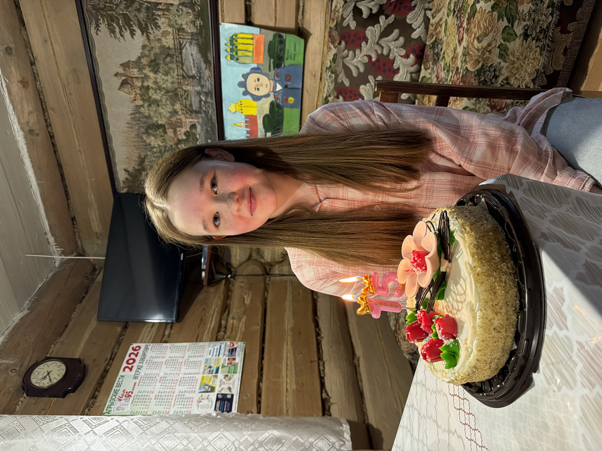 A young person is seated at a table with a birthday cake, presumably celebrating a 15th birthday given the candle numbers. The cake is decorated with artificial flowers and roses. The person has long brown hair, fair skin, and is wearing a pink checkered shirt.
The setting appears to be a rustic interior with wooden walls and a floral patterned curtain or upholstery in the background. On the wall behind the person is a landscape painting, a children's artwork, a calendar with the year 2026, a clock, and a flat-screen television. The location is Кадряково, Russia, as indicated on the calendar with "Агрызские вести" written in Russian. The table has a white patterned tablecloth.