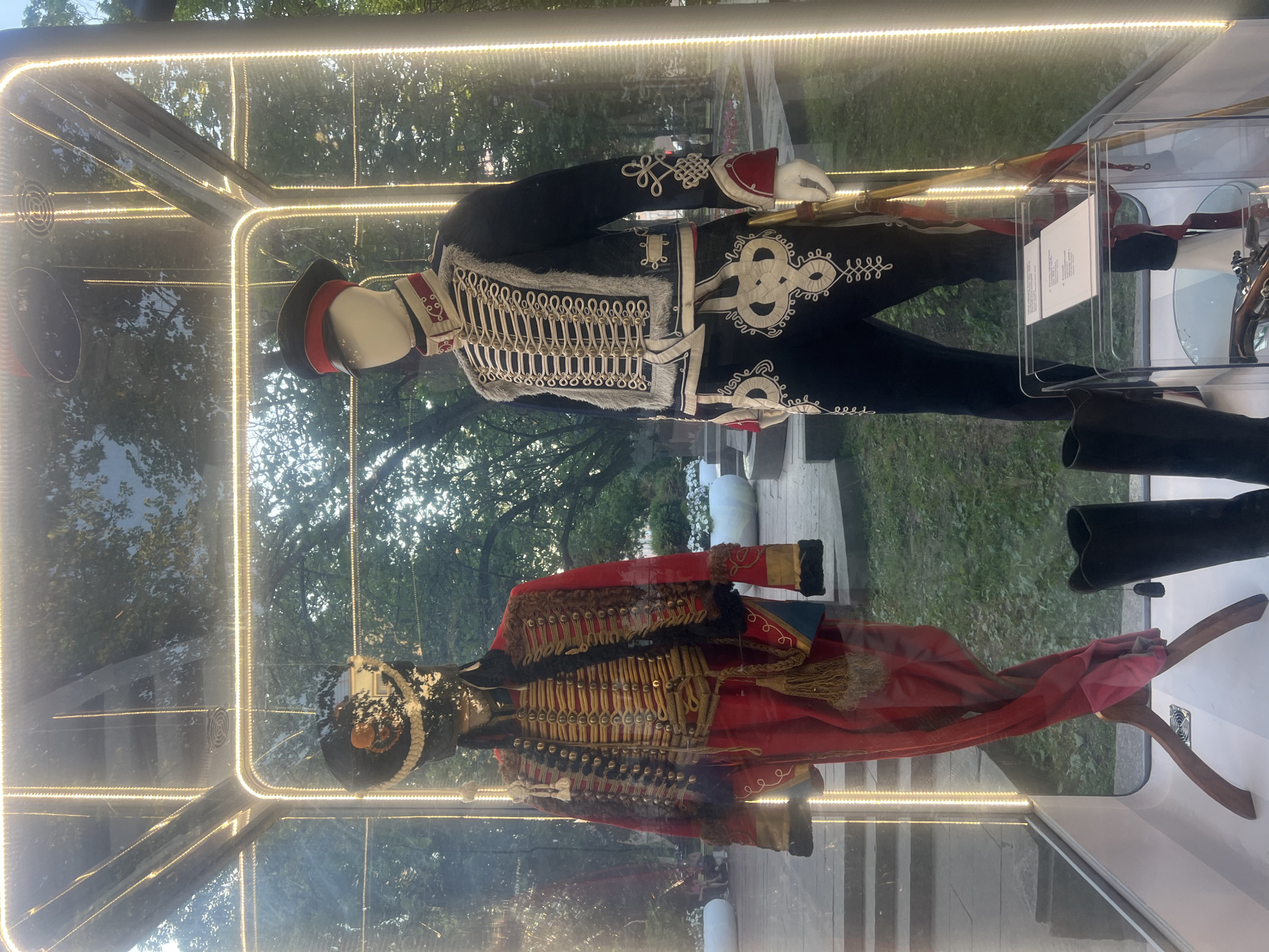 The image shows two mannequins dressed in historical military uniforms displayed inside a glass enclosure with integrated lighting. The enclosure is situated outdoors, with trees and greenery visible in the background, suggesting a park or a public space. One mannequin on the left wears a red uniform with intricate gold braiding and a fur-trimmed jacket, accompanied by a tall black boot. The mannequin on the right is dressed in a dark blue uniform with elaborate white braiding, a red-trimmed cap, and dark trousers, also with a tall black boot. A rifle is partially visible next to this mannequin. A placard is placed inside the enclosure in front of the right mannequin. The scene is likely a museum exhibit or a historical display in a public area of Moscow, Russia.