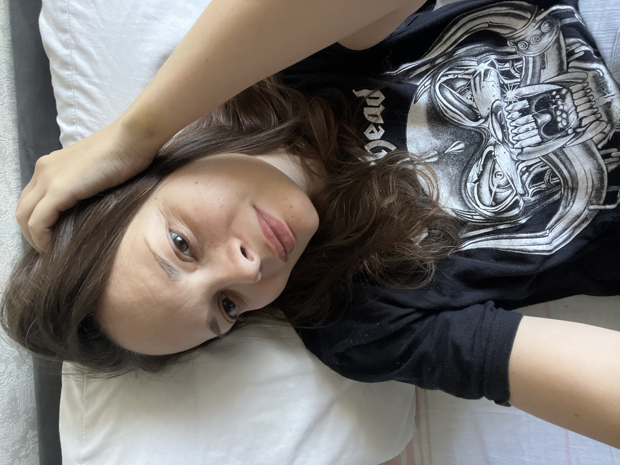 The image shows a young woman lying on her side in bed. She has brown hair and is wearing a black t-shirt with a graphic print of a skull. Her right hand is resting on her head, and her left arm is bent at the elbow. She is looking towards the camera with a slight smile. The background consists of a white pillow and a grey headboard. The overall scene suggests a casual, indoor setting, likely a bedroom. There is no specific information in the image that places it in Tolyatti, Russia.