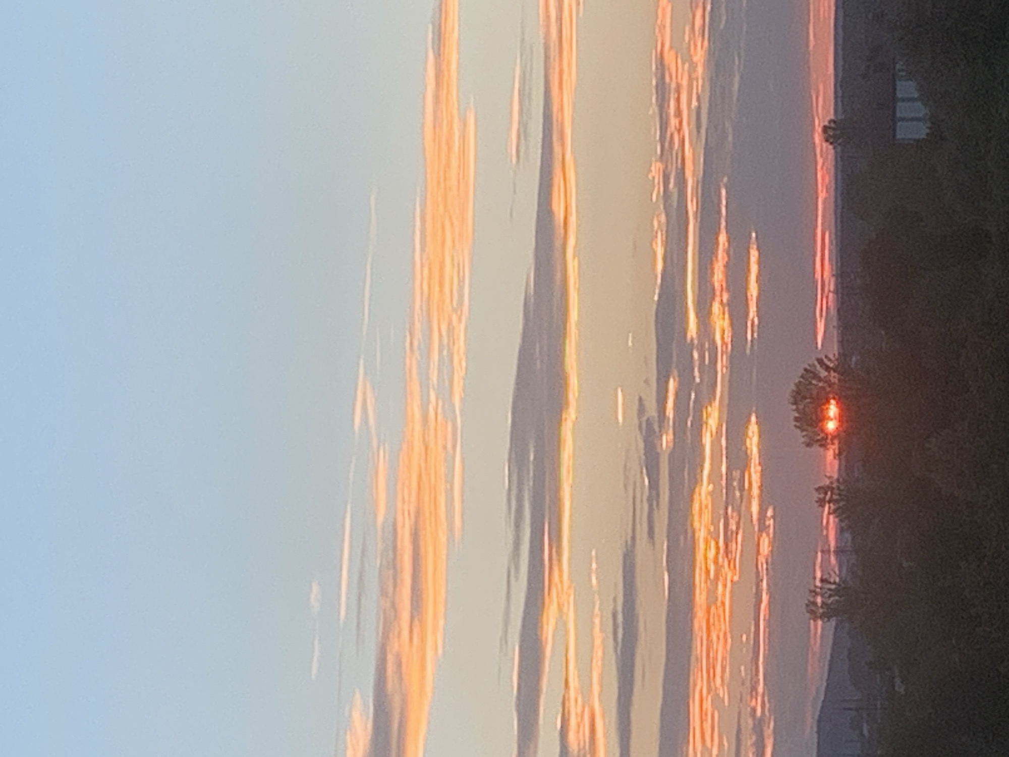 This image depicts a sunset over the city of Kolomyia, Ukraine. The sky is filled with horizontal bands of clouds, colored in shades of orange, pink, and purple, transitioning to a clear pale blue at the top. The sun is visible near the horizon, partially obscured by trees, casting a bright orange glow. The foreground is dominated by a silhouette of trees and vegetation. In the middle ground, a building with a light-colored facade is visible on the right. The horizon line shows rolling hills in the distance. The overall impression is one of a calm, natural landscape at dusk.