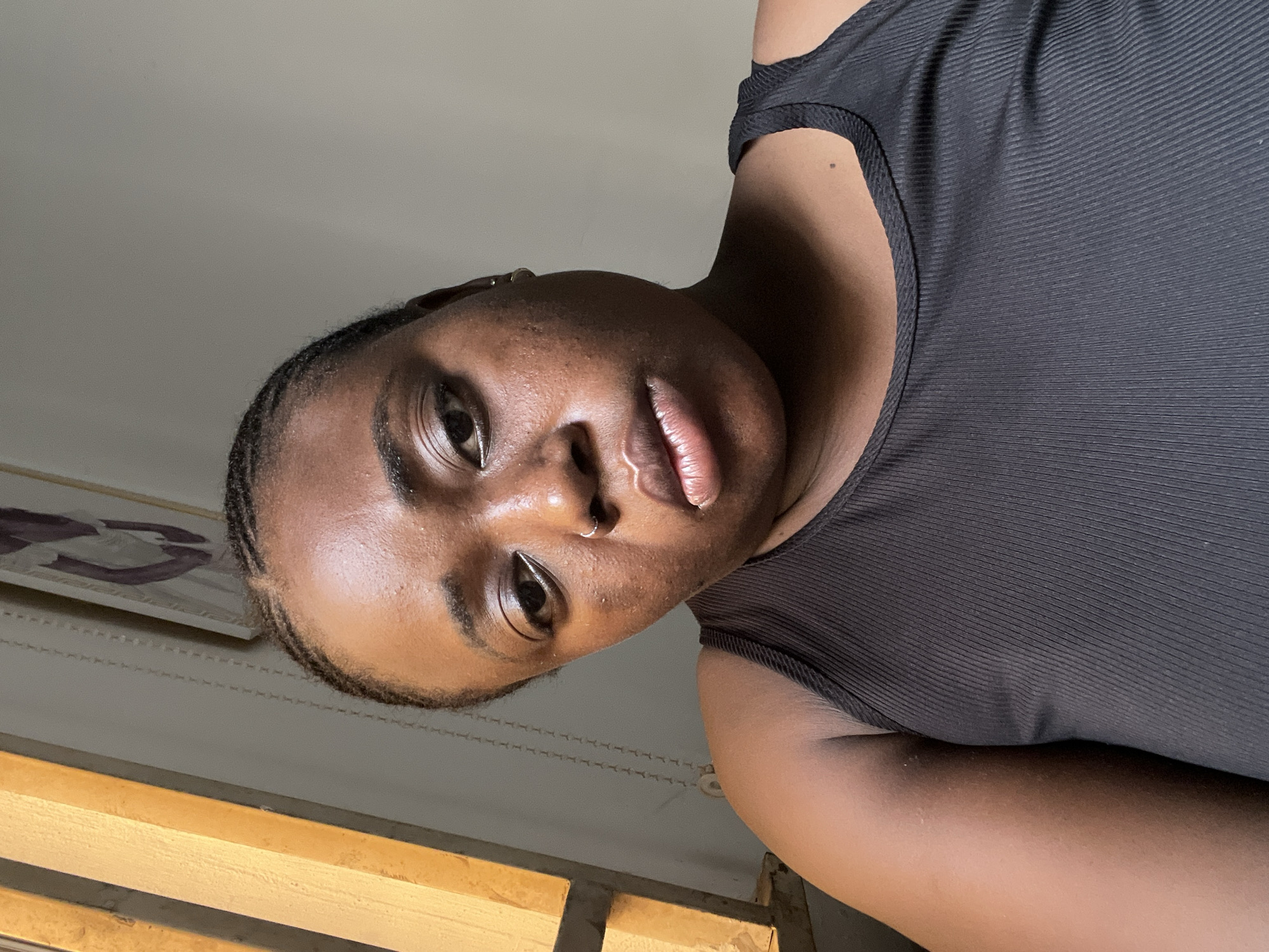 A young woman with dark skin, a nose ring, and braided hair is pictured in an eye-level shot taken from a low angle. She is wearing a black ribbed tank top. The background includes a white ceiling and part of a wooden structure, potentially a window frame. The scene is indoors, but no further details about the location in Abuja, Nigeria, are discernible.