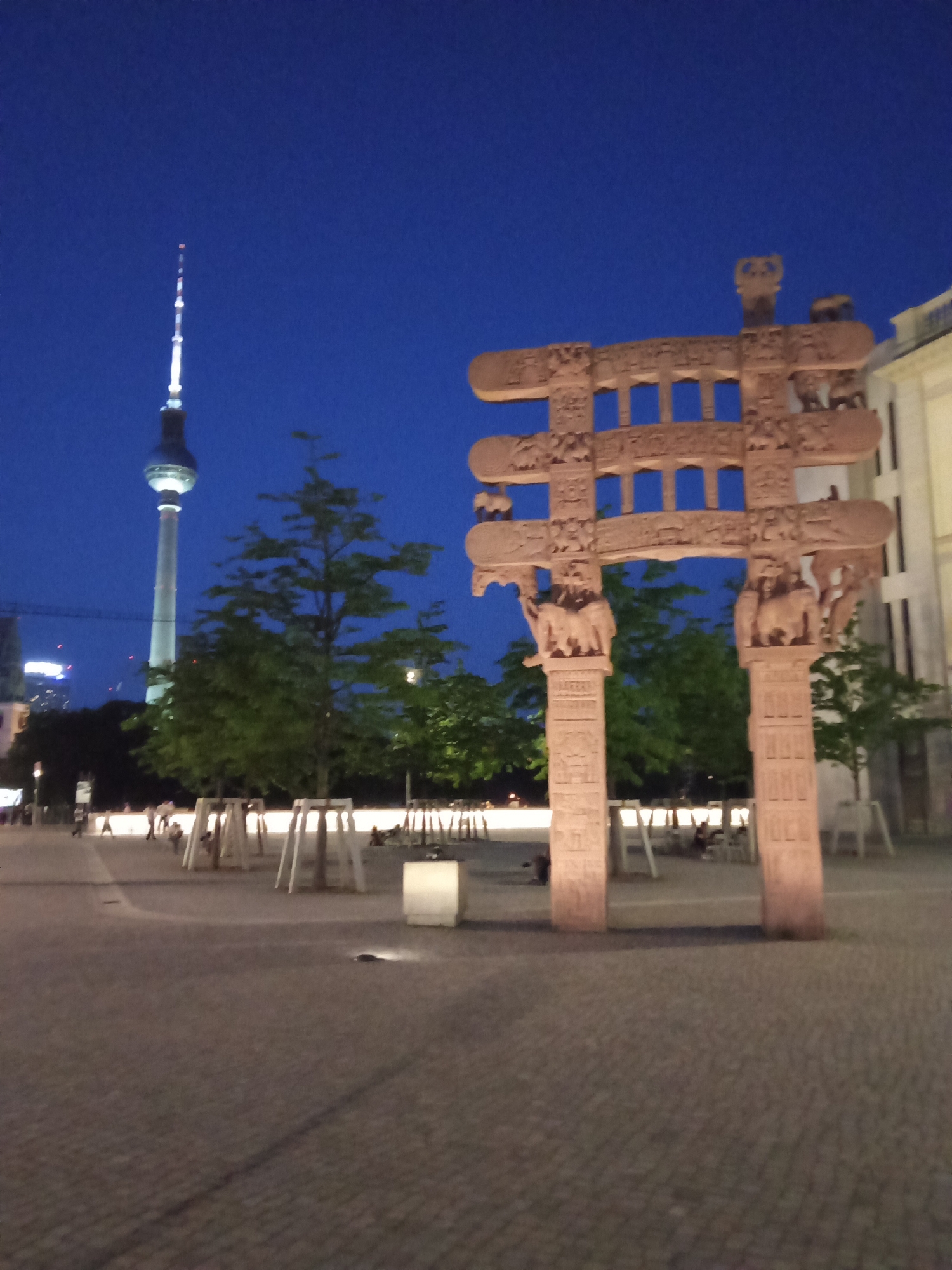The image shows the Brandenburg Gate in Berlin, Germany, at night. The gate is made of sandstone and features carvings of elephants and other animals. In the background is the Berlin TV Tower. The area around the gate is paved with cobblestones, and there are trees and benches in the vicinity. People are sitting on the benches and walking around the area. The sky is dark blue.
