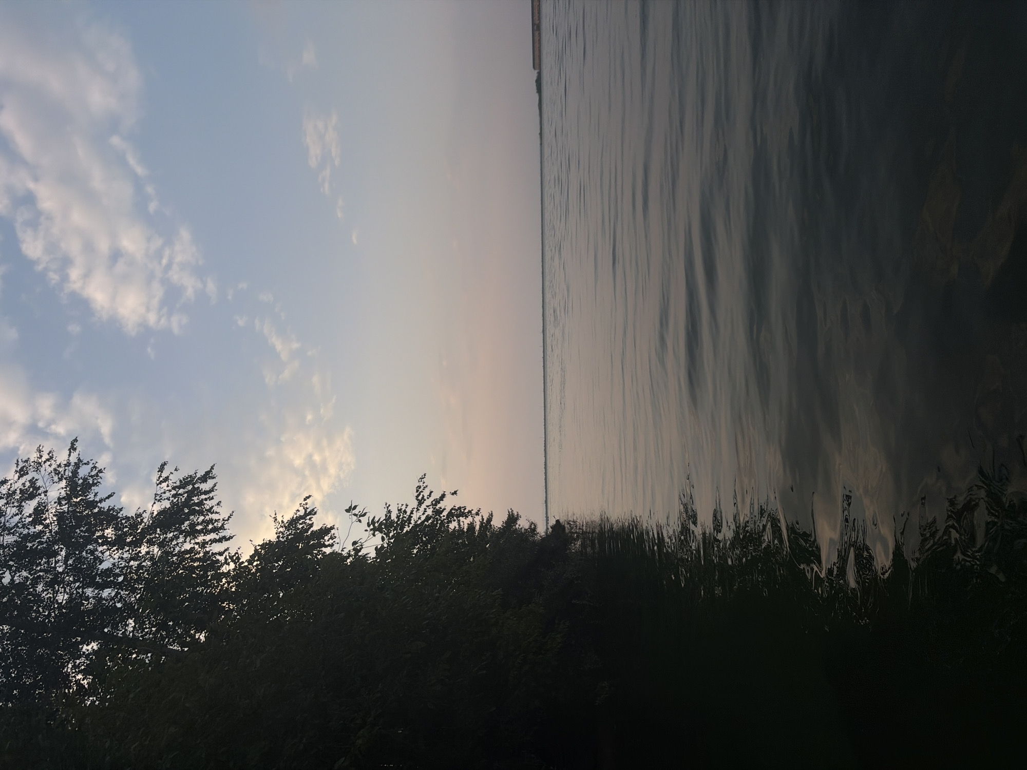 This is a photograph of the Kama River in Chaikovsky, Russia, taken from a low vantage point on or near the water, likely from a boat. The image captures the river during either sunrise or sunset, as indicated by the soft, diffused light and the muted colors of the sky.
On the left side of the frame, a dense line of dark green trees forms a natural embankment, their silhouettes sharp against the lighter sky. The trees appear lush and full, with some branches extending over the water. The water itself is calm, with gentle ripples reflecting the subtle hues of the sky. The right side of the image opens up to a wide expanse of the river, with a distant, low-lying shoreline visible. On this distant shore, there are some structures or landforms that appear to be man-made, possibly industrial buildings or a quay, characterized by their linear forms and darker tones.
The sky above is a gradient of soft blues and pinks, with scattered white clouds. The overall atmosphere of the photograph is serene and peaceful, with a focus on the natural beauty of the river and its surroundings, juxtaposed with the subtle presence of human development in the distance. There are no people or animals visible in the image.