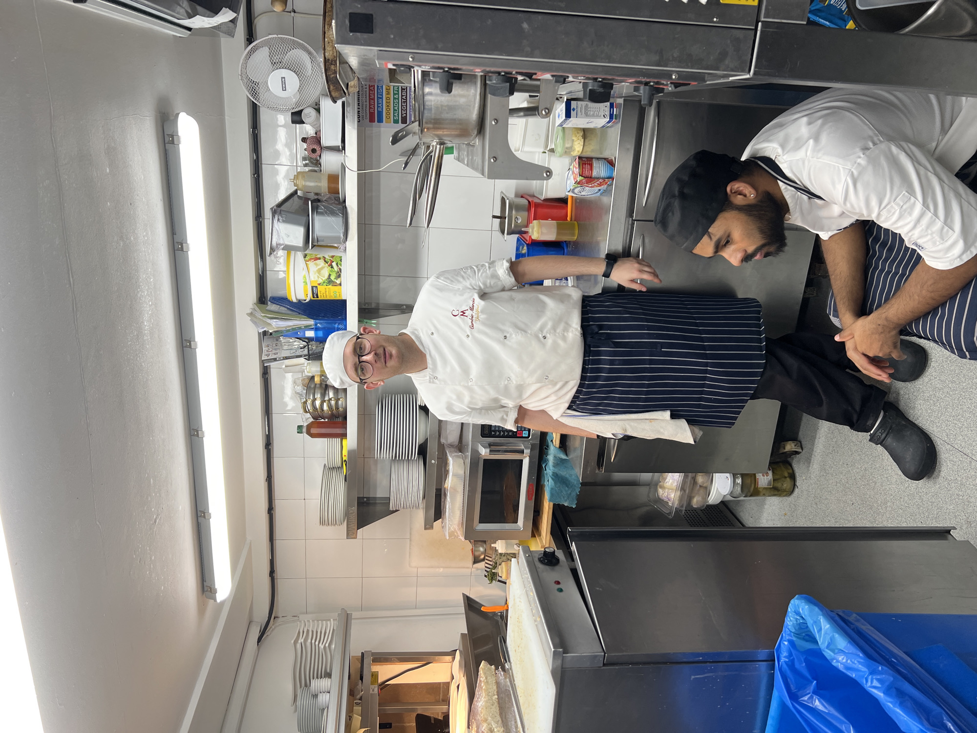 Two chefs are in a commercial kitchen. The chef on the left is standing and wearing a white chef's jacket, black trousers, and a blue and white striped apron. He has glasses and a chef's hat. The chef on the right is sitting and wearing a white chef's jacket and a black chef's hat. The kitchen appears to be well-equipped with stainless steel appliances, shelves stocked with plates and containers, and various cooking ingredients and utensils. The overall environment suggests a professional culinary setting. The location is identified as Guildford, United Kingdom.