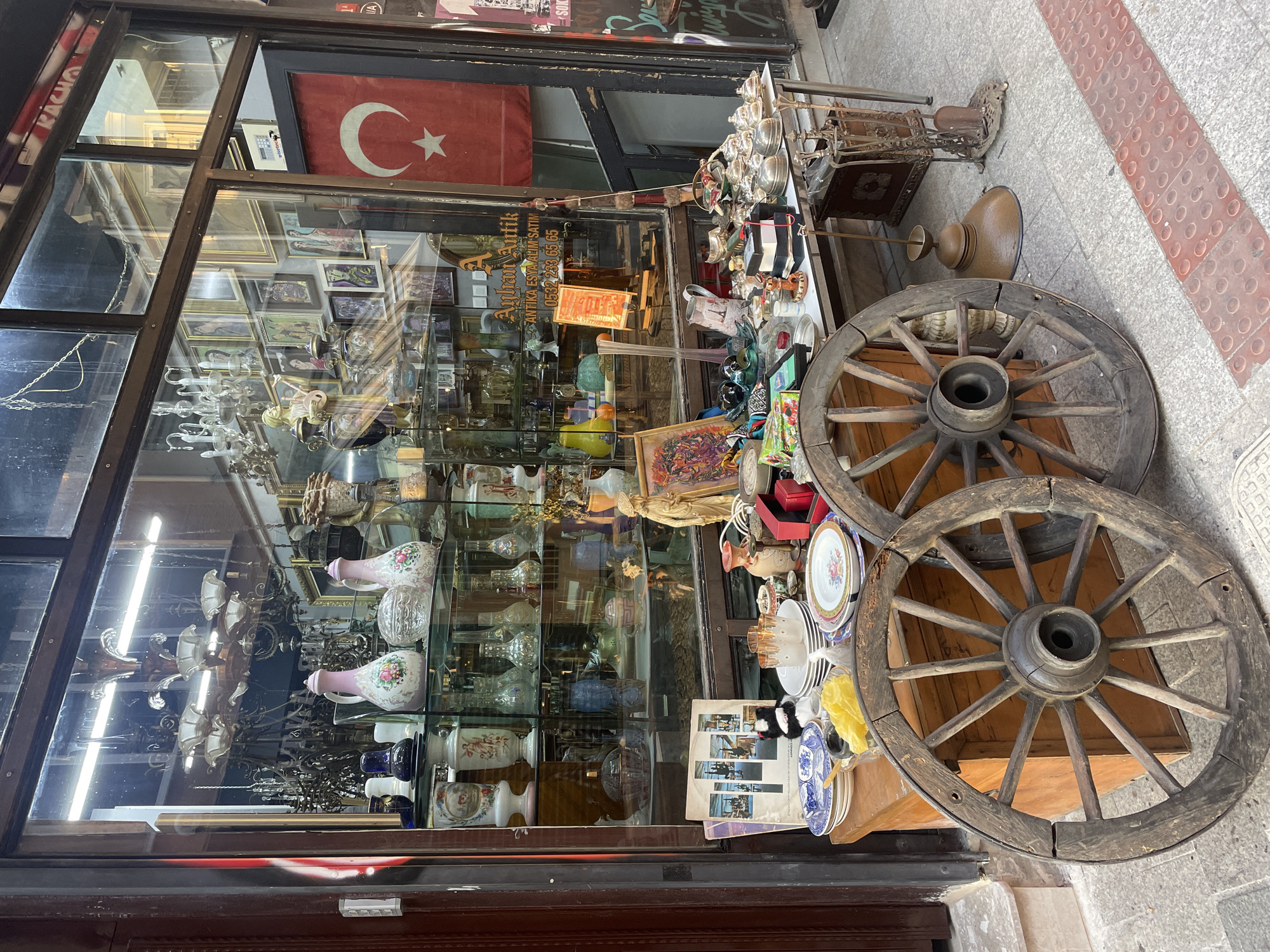 The image shows the exterior of an antique shop located in Kadıköy, Turkey. The shop's display window is filled with various antique items, including chandeliers, vases, plates, and decorative objects. In front of the shop, there are two large wooden wagon wheels placed on the sidewalk. A table in front of the window displays more items for sale, such as teapots, bowls, and plates. A Turkish flag hangs on the right side of the storefront. The overall scene depicts a busy street in a commercial area.