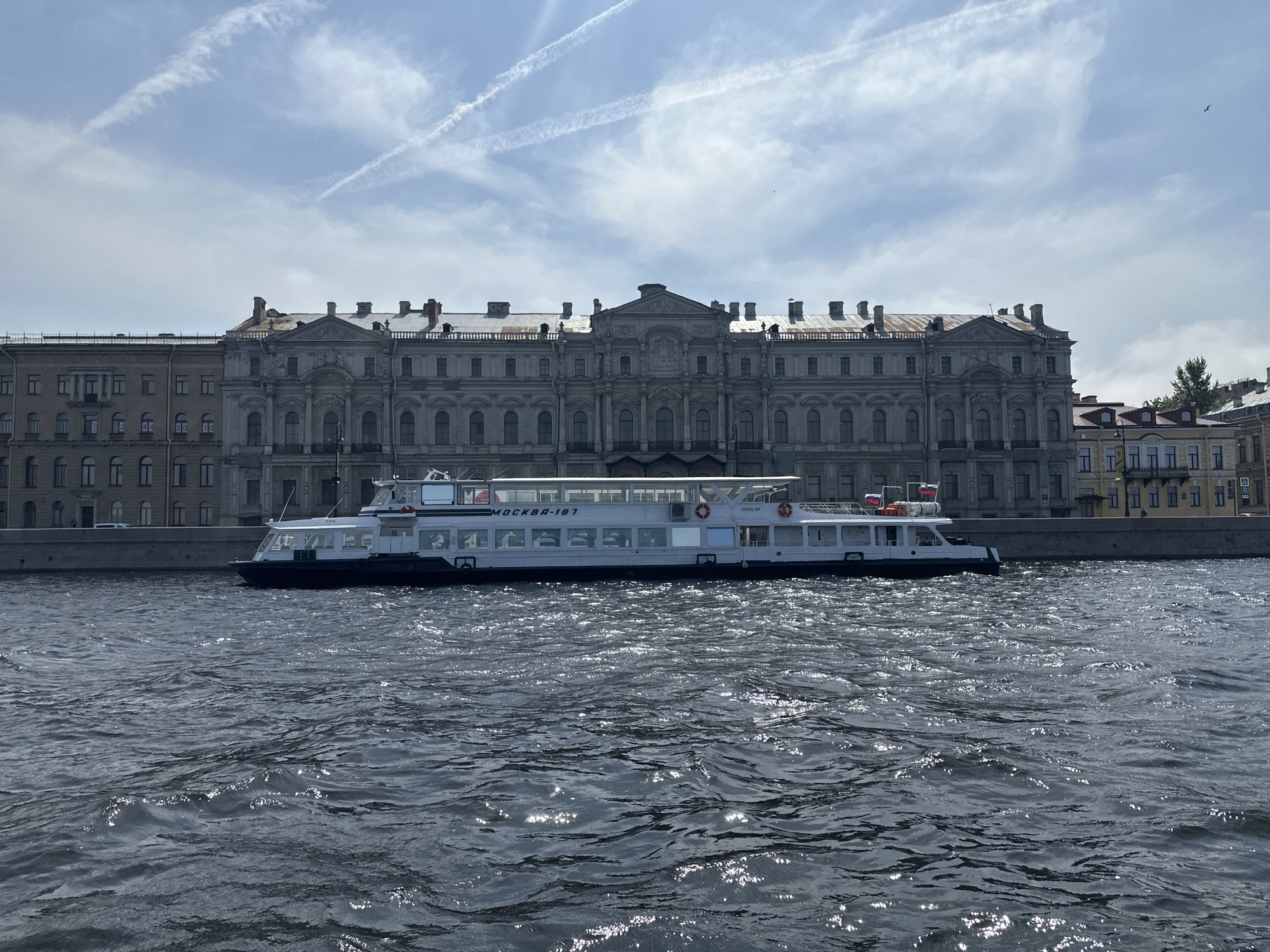 The image depicts a river scene in St. Petersburg, Russia, with a riverboat floating on the water in front of the Winter Palace. The water is dark with light reflecting off the ripples. The boat, "MOCKBA-187," is white and blue and situated parallel to the riverbank, obscuring a portion of the Winter Palace. The Winter Palace is a gray, ornate building with numerous windows and a pediment at the center. The sky is blue with thin contrails.