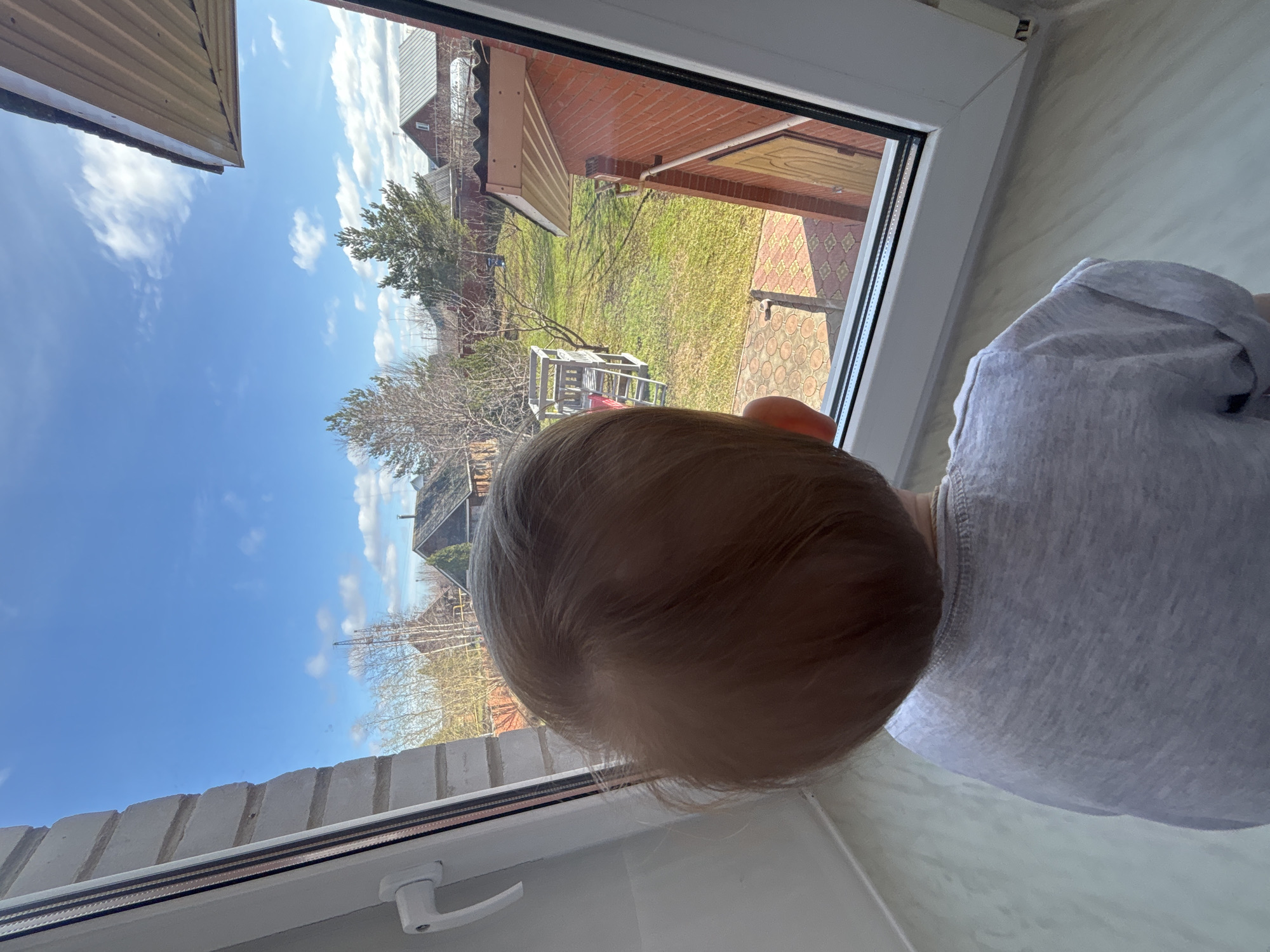 A child is looking out a window in Ферма, Russia. The window offers a view of a yard with grass, trees, and buildings. The sky is blue with some clouds. The child is wearing a light-colored shirt.