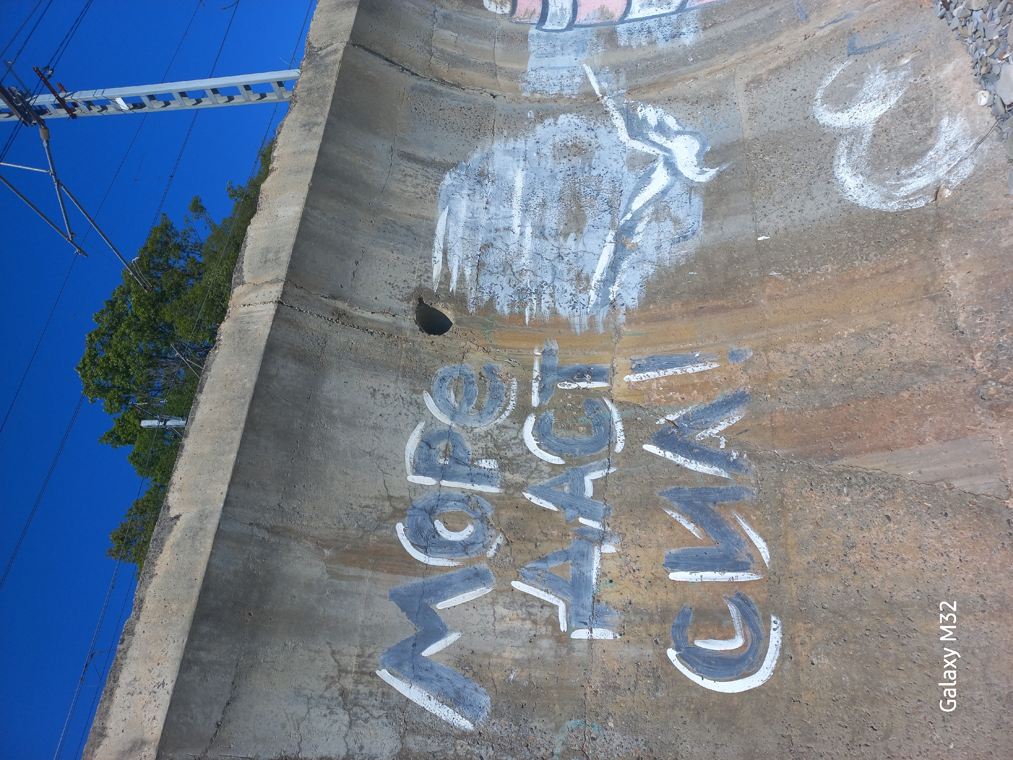 The image captures a curved, concrete wall with graffiti. The words "MORE PEACE GM" are spray-painted in gray with white outlines. Above these words, there is a white-painted figure, possibly an animal or abstract shape, that appears to have been partially covered or faded.
The concrete wall is aged and shows signs of wear and tear, with cracks and discoloration. At the top right of the wall, there is a glimpse of loose stones or gravel.
Above and to the left of the wall, the sky is visible, a clear, vibrant blue. Power lines and a metal structure are silhouetted against the sky. A tree with green foliage is visible behind the metal structure.
The camera used was a Galaxy M32, which is indicated in the lower right corner.
There is insufficient information to definitively locate this scene in Sochi, Russia, beyond the assumption made by the request.