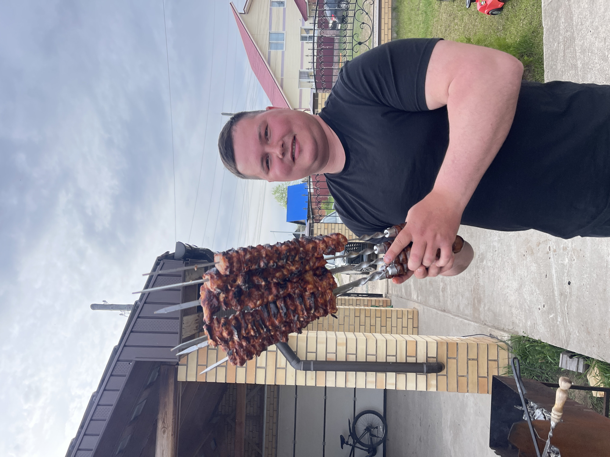 A man in a black t-shirt is holding skewers of cooked meat outdoors. The location appears to be a residential area in Chaikovsky, Russia. In the background, a building with a red roof and yellow siding is visible, along with a brick structure. A bicycle is leaning against the brick structure. A barbecue grill is visible near the bottom right of the image. The sky is overcast.
