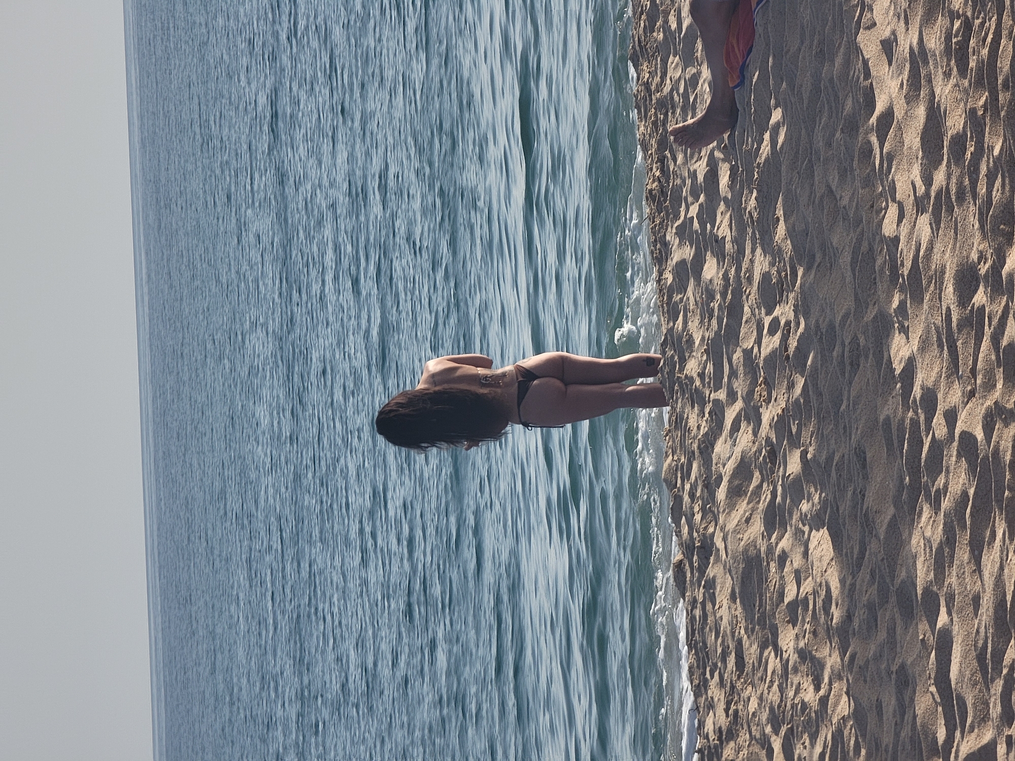 The image shows a woman in a black bikini standing at the edge of the water on a sandy beach. Her back is to the camera, and her long dark hair cascades down her back. The ocean is a deep blue, with gentle waves lapping at the shore. The beach is covered in sand, with some footprints visible. In the foreground, the legs of another person are visible, lying on a towel. The sky is a pale blue. The scene appears to be in Quarteira, Portugal, a popular tourist destination known for its beaches.