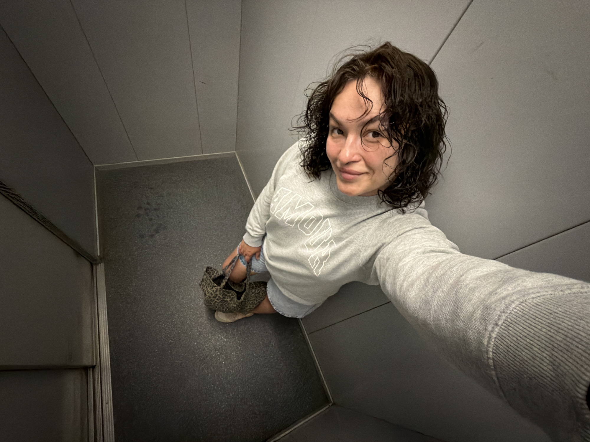 A young person with curly brown hair and a light gray sweatshirt is sitting on the floor inside an elevator. They are holding a leopard-print bag in their hands. The elevator walls are a neutral gray color, and the floor is dark gray with a textured pattern. The lighting is dim, casting shadows on the walls and floor. The image appears to be a selfie taken from a high angle. The location of Chaikovsky, Russia, is not discernible from the image.