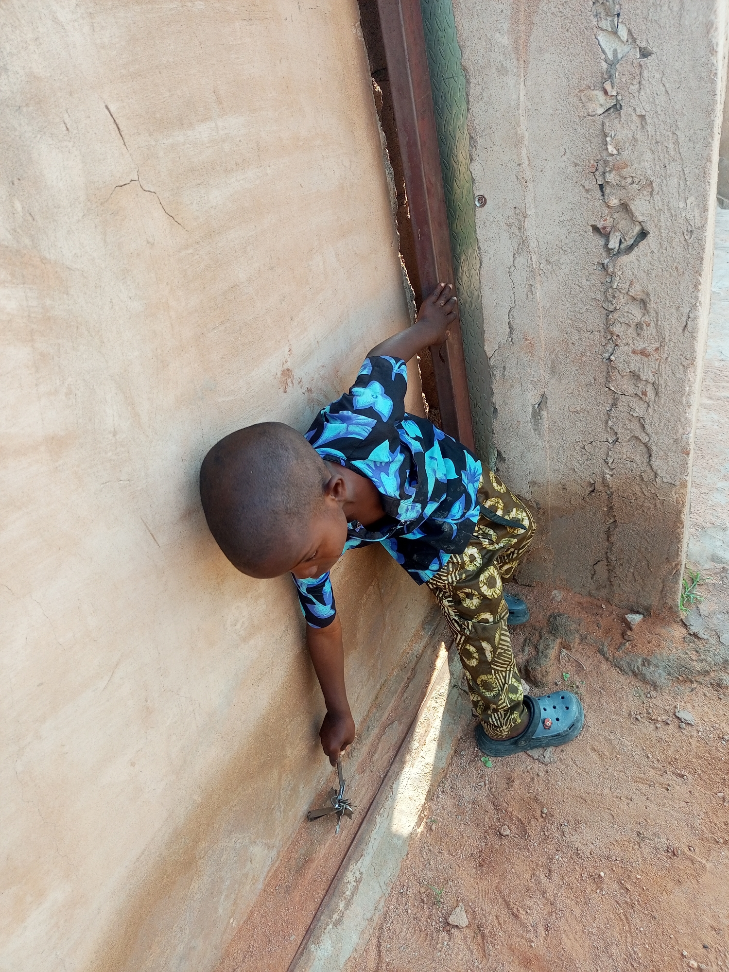 A young boy, appearing to be of African descent, stands near a weathered wall and a metal gate. He is wearing a black and blue patterned shirt and patterned pants, along with black clogs. The boy is holding a set of keys and appears to be reaching towards the wall or gate. The ground is covered in dirt and small stones. The environment suggests an outdoor setting, possibly in a residential area of Tempure, Nigeria. The wall shows signs of age and damage, with cracks and chipped plaster. The metal gate is partially visible. Sunlight casts shadows, indicating it is daytime.