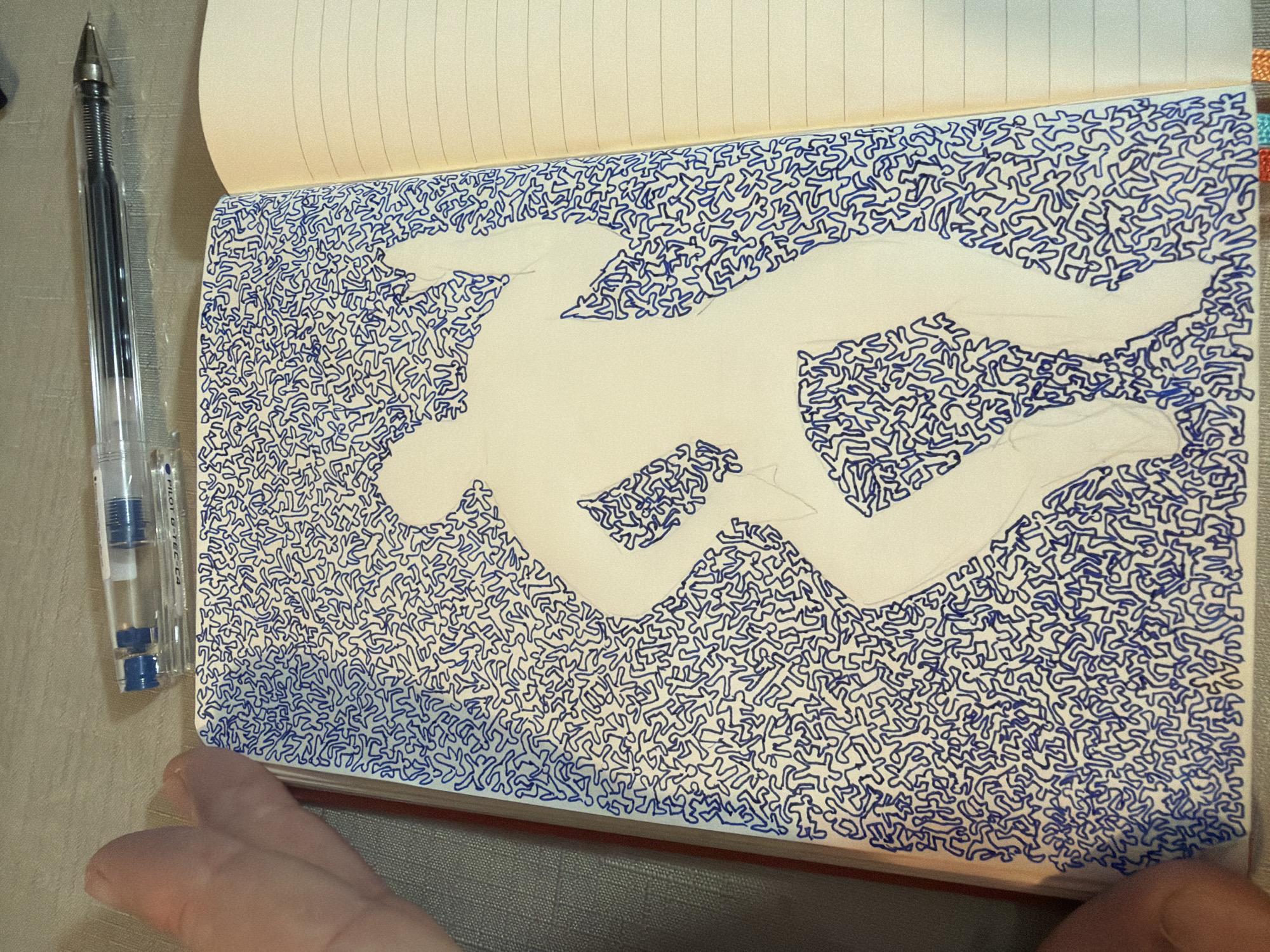 The image shows an open notebook with a drawing on its left page. The drawing is a silhouette of a dancing person, filled with a pattern of small, blue, abstract human-like figures. The background of the notebook page is a light cream color. To the left of the notebook, a hand with a thumb extended is visible, seemingly holding the notebook open. Above the notebook, a clear plastic pen with a black ink cartridge is resting horizontally. The notebook is placed on a textured surface that appears to be a tablecloth or a similar fabric. The right page of the notebook is blank and lined. There is no visible information or context to indicate the location in the city of Even Yehuda, Israel.