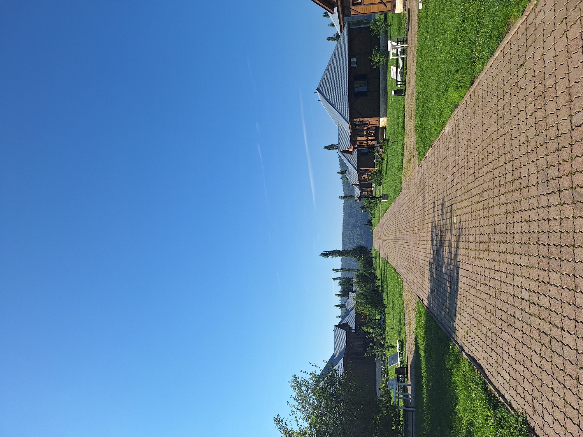 The image depicts a paved pathway leading through a village of wooden cabins in Atamanovka, Russia. The sky is a clear, bright blue. On either side of the path, well-maintained green lawns are dotted with small trees and bushes. Several cabins, constructed with dark wood and featuring corrugated metal roofs, are visible along the right side of the pathway. Each cabin appears to be a detached dwelling. On the left side, more cabins are partially obscured by lush green trees and foliage. White lounge chairs and a small table are present on the grass near the cabins on the left. Towards the horizon, a densely forested hillside meets the sky. The scene suggests a tranquil, resort-like environment, likely a tourist accommodation or a residential village.