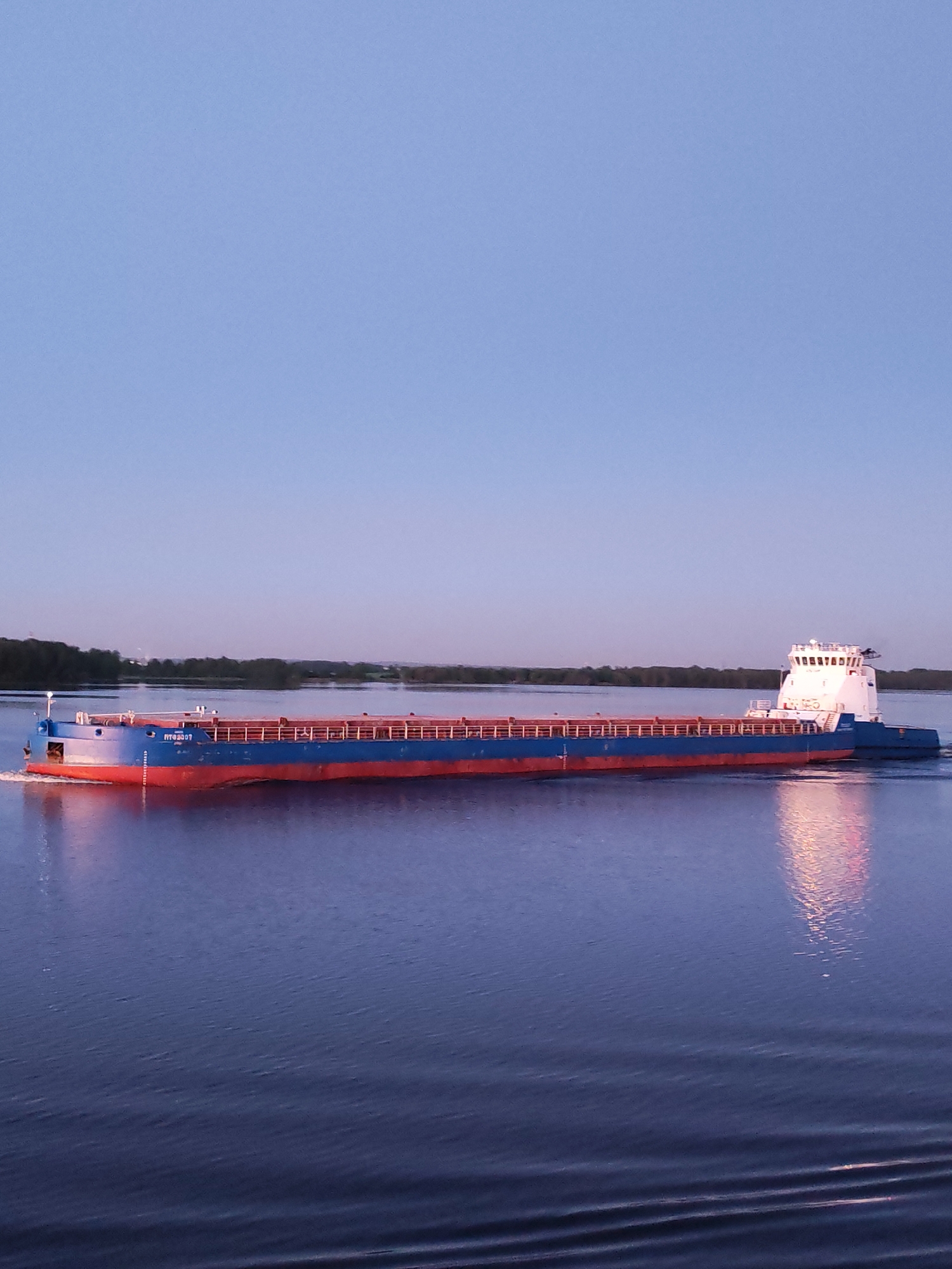An elongated river cargo vessel, predominantly dark blue with a red hull below the waterline, traverses a wide body of water from left to right under a clear, light blue sky. The vessel features a flat deck with reddish-brown cargo hold covers and blue railings. White Cyrillic text "РТС2007" (RTS2007) and "СВЕРДЛОВСКЪ" (SVERDLOVSK) are visible on its bow. A white pushboat with a multi-windowed superstructure, identified as "ОКА-3205" (OKA-3205), is integrated at the stern, propelling the cargo section. Warm, bright reflections from the pushboat's lights or structure shimmer on the dark blue water surface, creating prominent vertical light streaks. Ripples extend behind the moving vessel, indicating its forward momentum. The sky above is a clear, light blue, gradually lightening towards the horizon. A distant, low-lying shoreline covered in dark green trees is visible across the water. The scene takes place in the city of Unknown, Unknown.