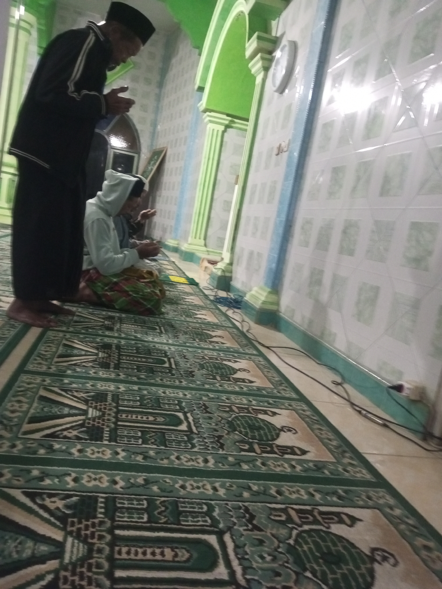 The image depicts an interior scene, likely a mosque, in Andongsili, Indonesia. In the foreground, a man wearing a black cap and black robe stands, facing left with his hands clasped, engaged in prayer. To his right and slightly in front, a younger person in a light blue hooded sweatshirt and a colorful sarong kneels on a patterned rug, also appearing to be praying. The environment features walls tiled in a light color with a repeating geometric pattern, and green architectural elements, including columns and an arched ceiling. A clock is visible on the wall. The floor is covered with prayer rugs featuring intricate green and cream designs. Cables are seen on the floor. The lighting is dim, with light reflecting off the tiled walls.
