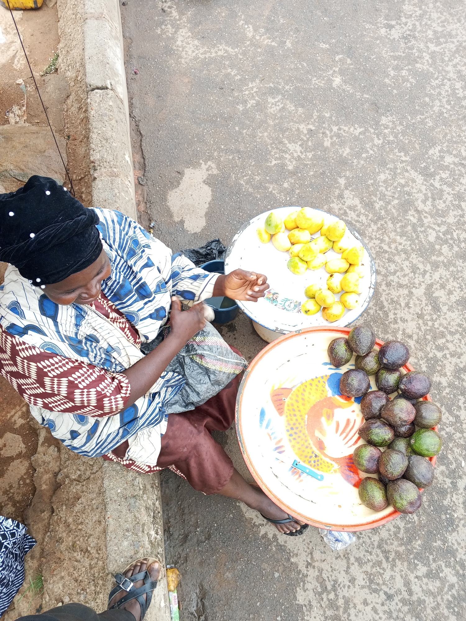 A woman sits on the edge of a sidewalk in Gwong, Nigeria, selling fruit. She wears a black head covering with white pearls, a patterned brown, white, and blue long-sleeved shirt, and brown pants. To her right are two round trays; the tray closest to her contains avocados, and the further tray contains mangoes. She is holding a clear plastic bag. A man's foot is partially visible at the bottom of the frame. The ground is concrete.