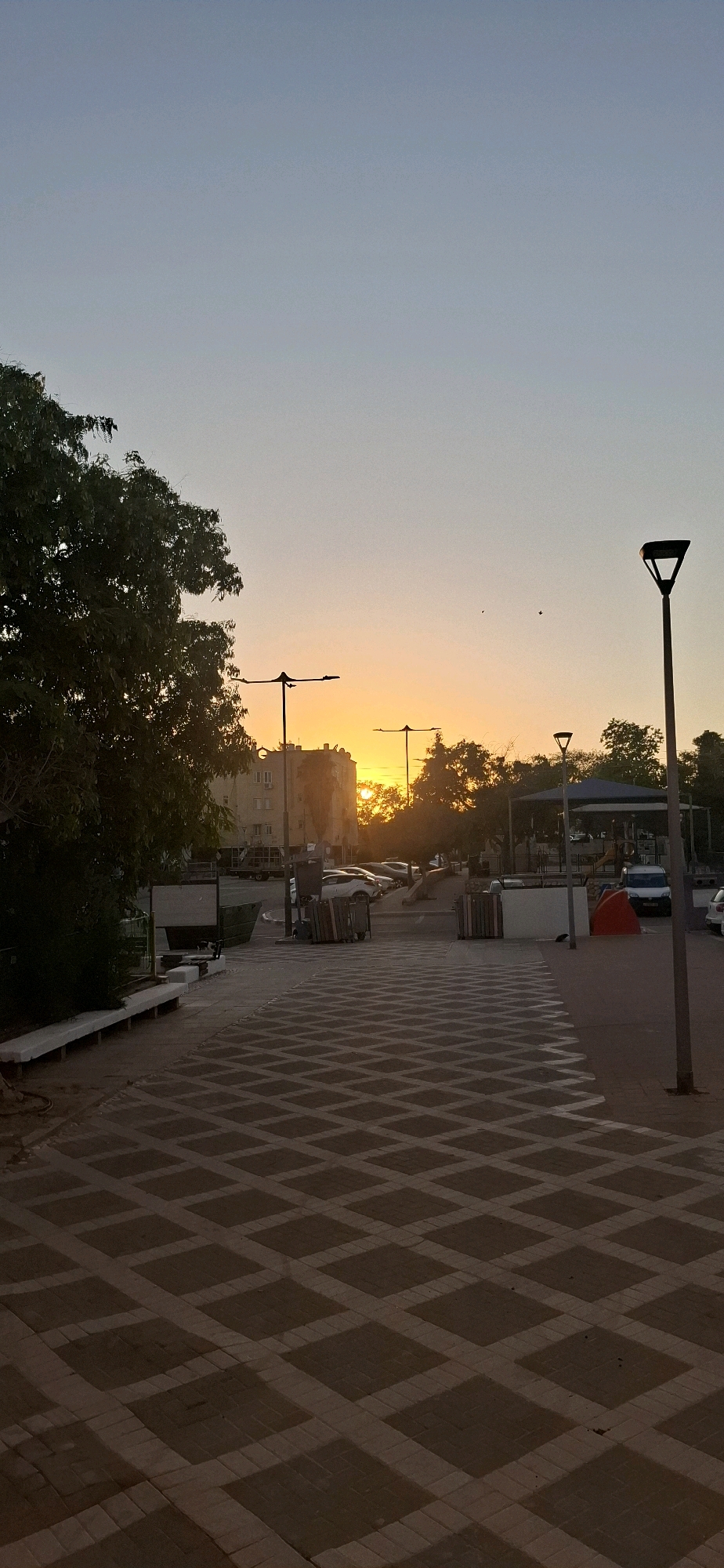 The image depicts a street scene in Yerocham, Israel, during sunset. The sky displays a gradient of pale blue to orange, indicating the sun is low on the horizon. In the background, a building with multiple windows is visible, partially obscured by trees. Several streetlights are present, one of which is prominent in the foreground on the right. The ground is paved with a pattern of light and dark colored diamond shapes. Cars are parked in the middle ground. A large tree with dark green foliage is on the left side of the image. The overall impression is a quiet urban environment at dusk.