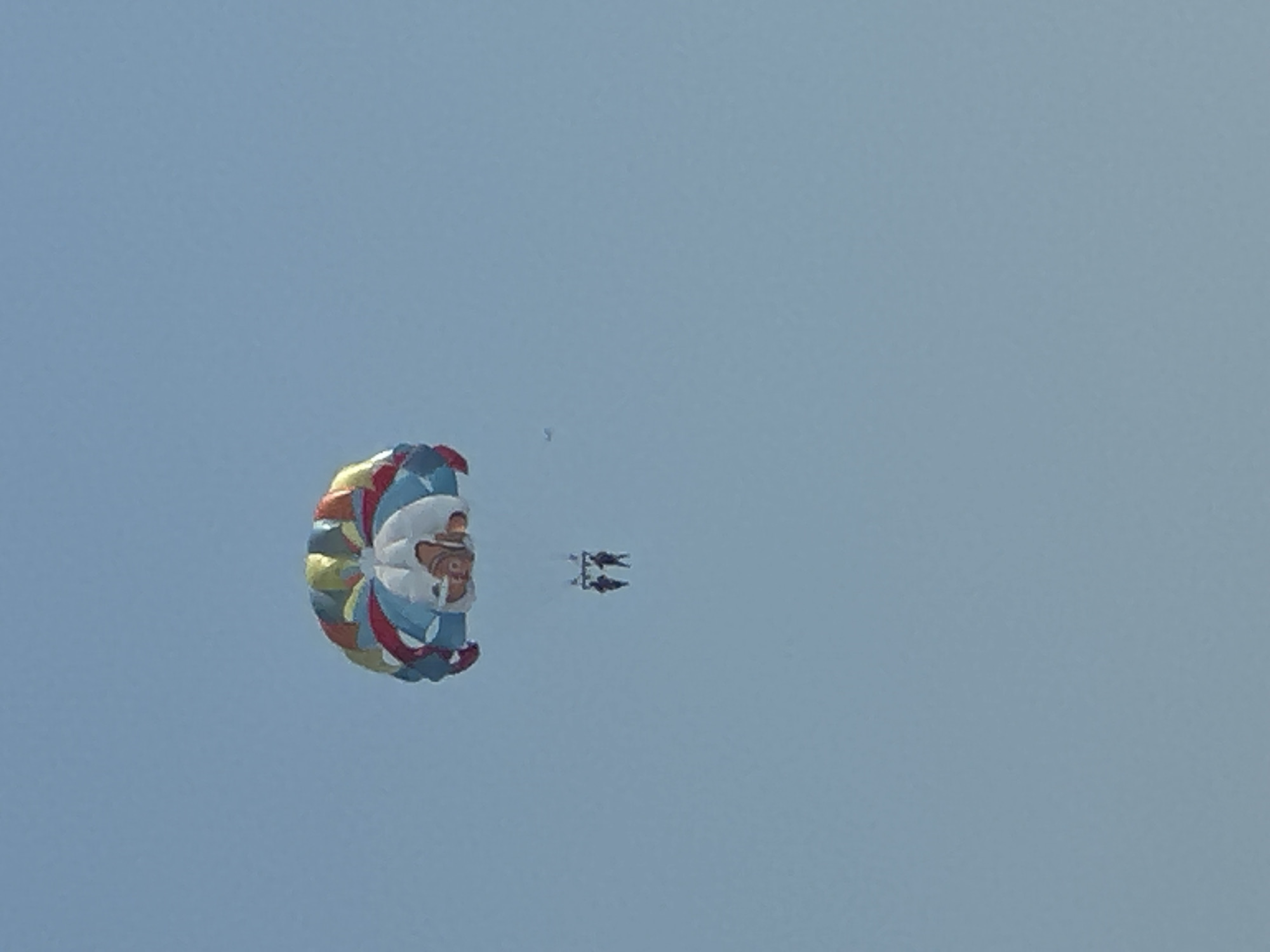 The image captures a clear blue sky, typical of a sunny day. A colorful parasail, adorned with a character resembling Nemo from the movie "Finding Nemo", is visible in the upper portion of the frame. Suspended beneath the parasail are two figures, presumably engaged in parasailing. Their dark silhouettes stand out against the bright sky, indicating they are at a considerable height. The scene is devoid of any urban landscape or specific landmarks that would pinpoint a location within the city of Boreti, Montenegro. The overall impression is one of recreational activity taking place in an open, aerial environment.