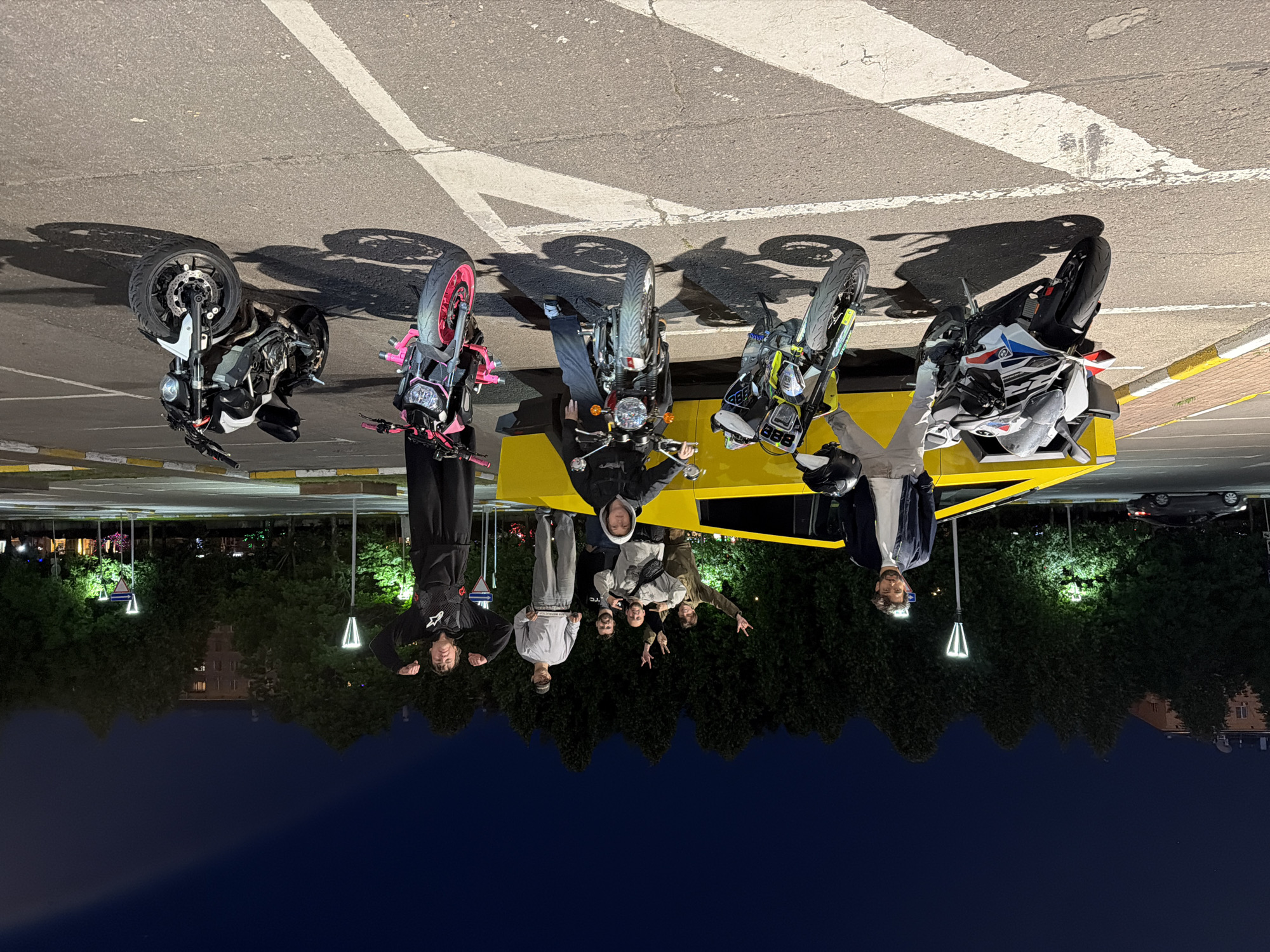 This image captures a nighttime gathering of at least seven individuals, predominantly young men, with their motorcycles in what appears to be an urban parking lot or street in Moscow, Russia. The scene is brightly lit by modern streetlights, casting shadows from the vehicles and people onto the asphalt ground, which features clear white road markings.
In the center, the group stands around five motorcycles and a bright yellow car. From left to right, the motorcycles include a black and white sportbike, a pink-accented motorcycle with matching pink wheels and grips, another dark sportbike, a yellow and black supermoto-style bike bearing the number "888" on its front, and a white, blue, and red sportbike. The yellow car provides a vibrant backdrop for the central figures.
The people are dressed in casual attire, including jackets, hoodies, and tracksuits. One individual on the far left is seen flexing, wearing a black tracksuit with a white star logo. Another person's light grey pants show partially visible "SUPREME" text. Further to the right, two individuals gesture peace signs towards the camera. The overall mood appears casual and social, suggesting a meet-up of motorcycle enthusiasts.
The background consists of a dense line of trees silhouetted against the dark night sky, with glimpses of distant buildings. The clear night sky and artificial lighting confirm the nighttime setting. No specific weather conditions are indicated other than clear skies.