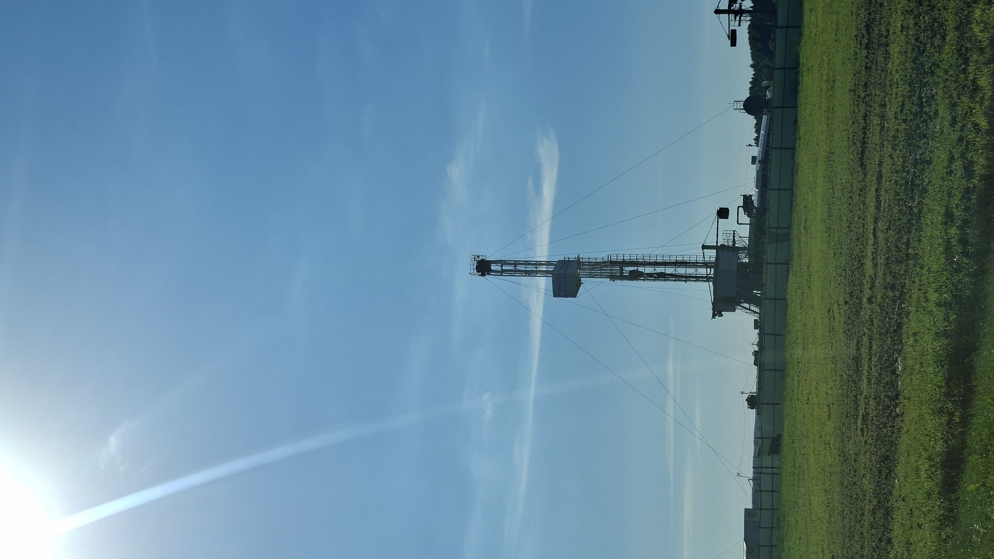 This is a vertical, eye-level, outdoor photograph of a drilling rig. The rig, a tall, metallic structure, is silhouetted against a bright blue sky with some wispy clouds and a strong sun flare in the upper left corner. The rig is surrounded by a fence, behind which are various pieces of industrial equipment and some trees in the distance. In the foreground is a green, grassy field. The scene appears to be in an industrial area or a rural setting where drilling operations are taking place, likely within the Бешанковіцкі сельскі Савет in Belarus, given the context. There are no people visible in the image.