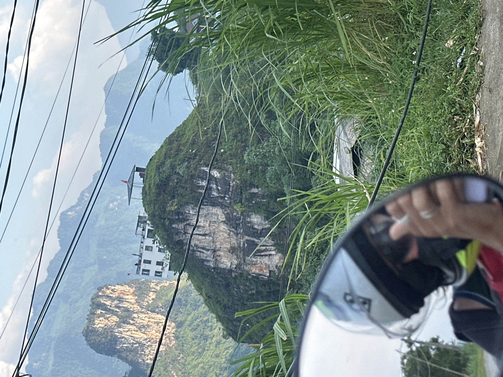 From the perspective of a motorcycle, the photo shows a mountain with a white building and lookout tower on top. The mountain is covered in greenery and has a distinctive rocky face. Power lines run across the scene. The motorcycle's side mirror reflects an image of the rider and surrounding foliage. The location is Đường Thượng Commune, Vietnam.