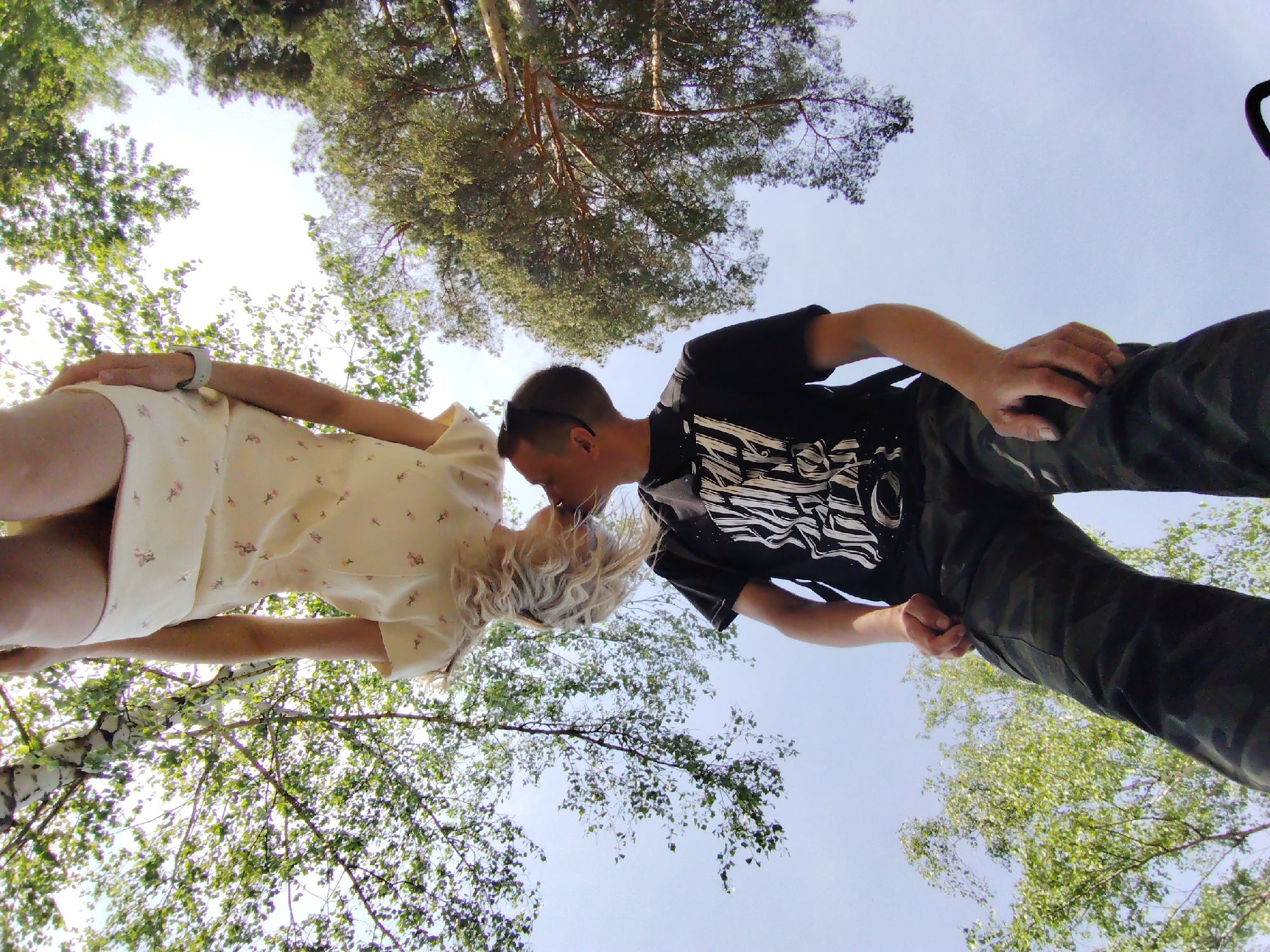 A low-angle shot depicts a couple, a woman on the left and a man on the right, seemingly kissing. The woman wears a light-colored dress with floral print, and the man is in a black t-shirt with white graphics and dark pants. The background is a view looking up into trees and sky, suggesting the setting is outdoors, possibly a park or wooded area. The trees are dense with green foliage. While the specific location in Chaikovsky, Russia cannot be forensically determined without further information, the presence of birch trees and pine trees is consistent with the natural environment of that region. The couple's interaction is the central focus, with the surrounding environment providing context.
