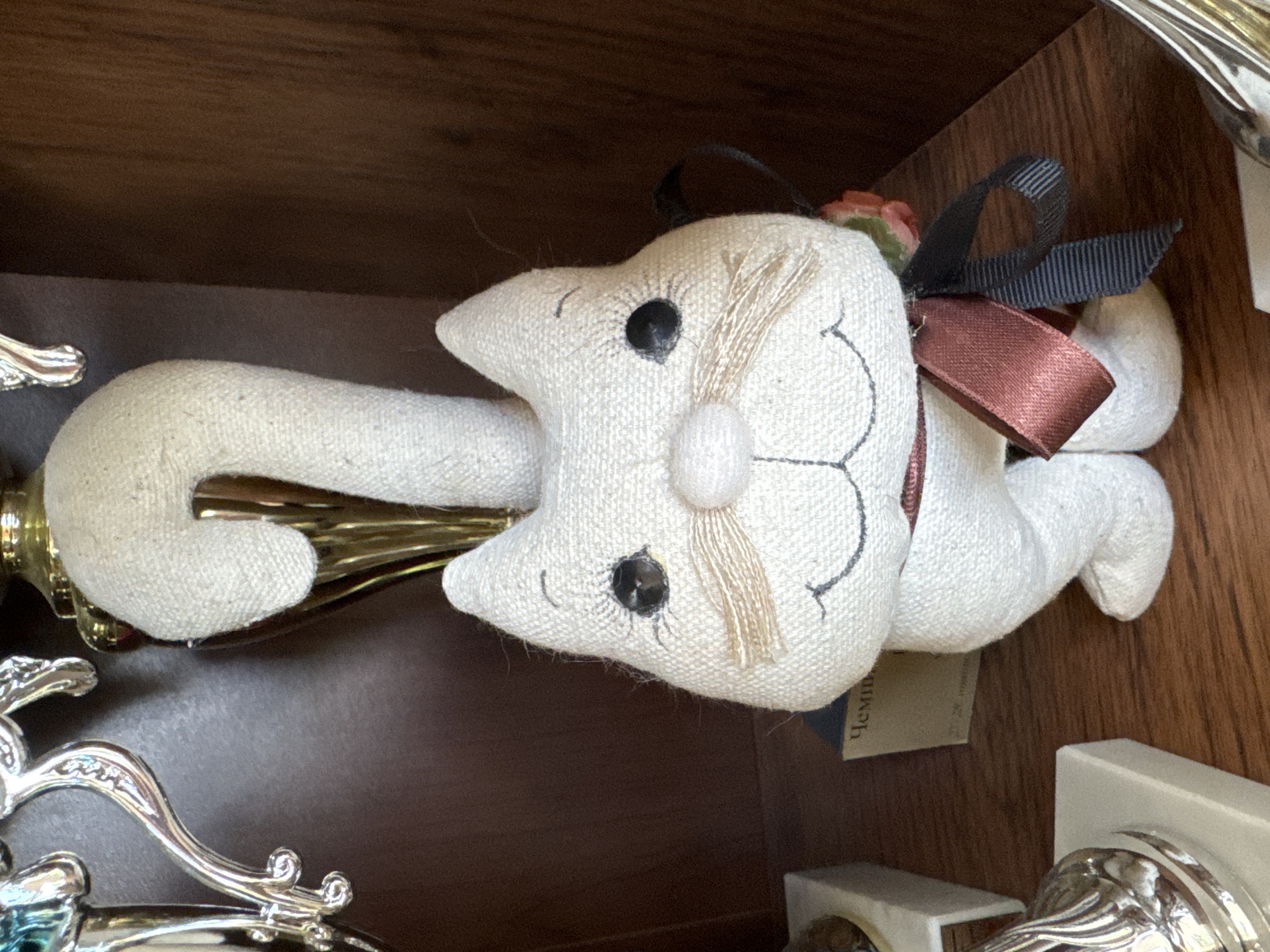 The image shows a stuffed cat on a shelf, likely in Steklyannyi, Russia, given the context. The cat is made of white, textured fabric and has black button eyes and embroidered features. It is wearing a dark red ribbon tied in a bow around its neck, with a black and gray striped ribbon also attached. Behind the cat, a golden trophy or vase is visible, and to the left, a silver trophy. The shelf is made of dark wood, and a small, pale blue and beige box with Cyrillic lettering is partially visible behind the cat's legs. The overall lighting suggests an indoor setting.