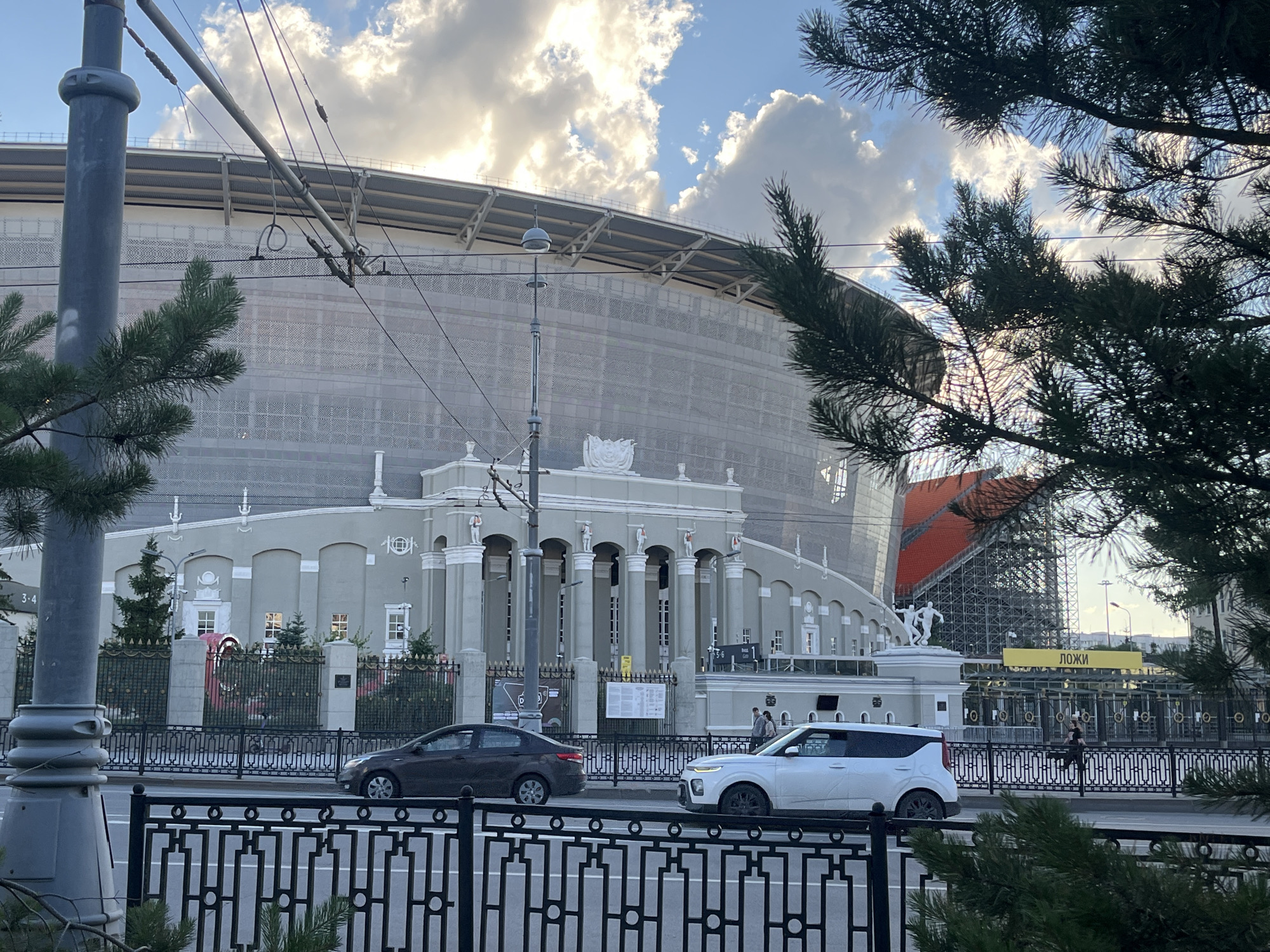 This media file captures an outdoor urban scene in Yekaterinburg, Russia, likely during the late afternoon or early evening, given the warm lighting and cloud formations. The sky is partly cloudy with bright sunbeams breaking through, suggesting fair weather.
The focal point of the image is a large, imposing building with a curved facade, possibly a stadium or a large public venue. The building is partially covered in a light-colored mesh, suggesting ongoing construction or renovation. In front of this structure, a more ornate, classical-style building with columns and decorative elements is visible.
In the foreground, a street runs horizontally, with two cars driving on it: a dark sedan and a white SUV. A black metal fence separates the street from the sidewalk and the building complex. Trees, specifically pine trees with their needles, frame the top and sides of the image, adding a natural element to the urban landscape.
There are a few people visible on the sidewalk, appearing to be pedestrians. One section of the building has a prominent yellow sign with Cyrillic text that reads "ЛОЖИ", which translates to "Boxes" or "Loges". The overall mood is that of a bustling city with significant architectural structures and ongoing activity.