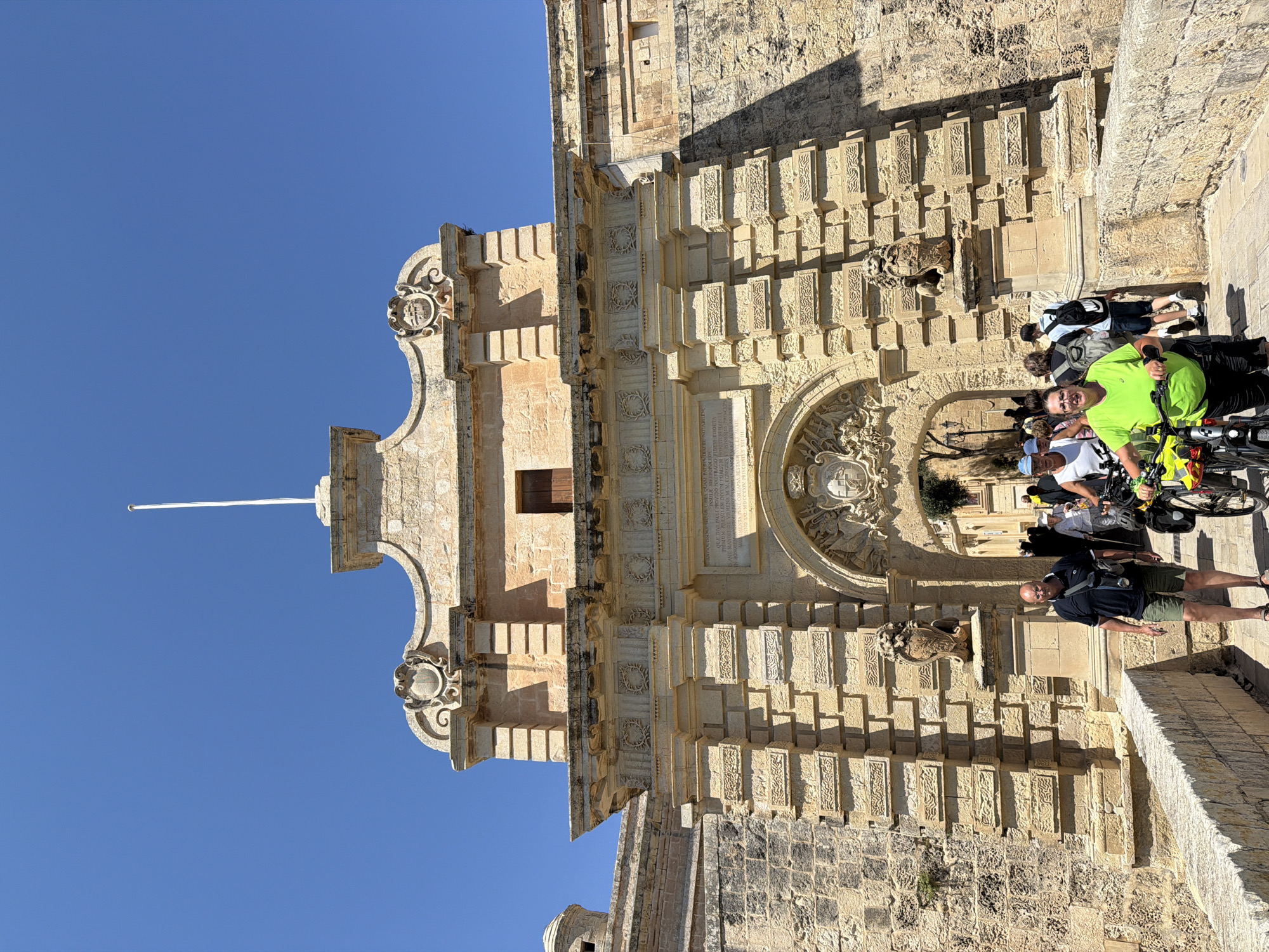 The image depicts a section of a sandstone building under a clear blue sky. The building has ornate detailing, including rectangular protrusions and decorative carvings. A group of people, including individuals on bicycles, are situated near the building. The location is in the city of Unknown, Unknown.