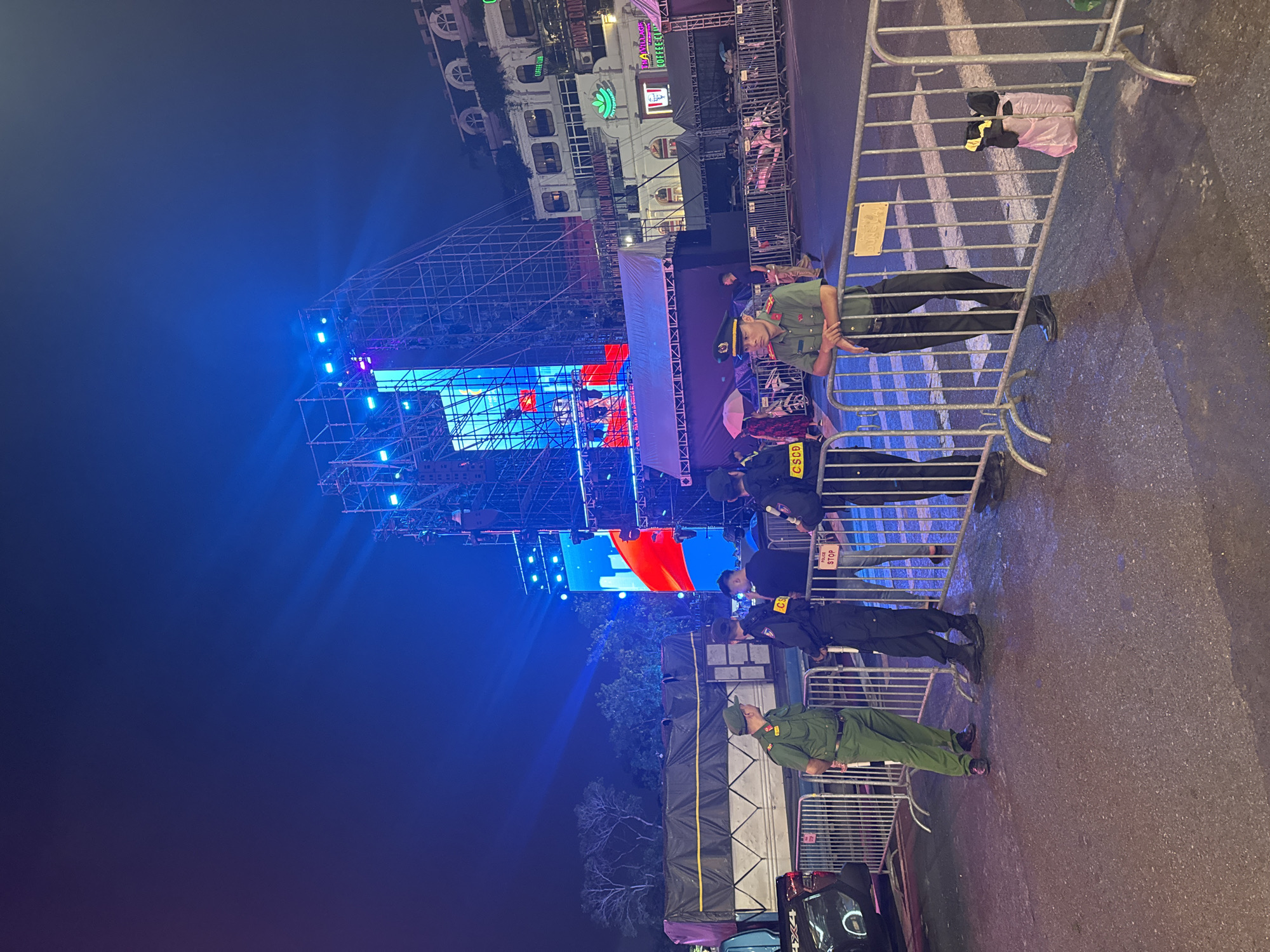 The image depicts an outdoor night scene in Hanoi, Vietnam, likely at a public event. A large stage structure, illuminated by blue and white lights, dominates the background. Scaffolding surrounds the stage, indicating it is under construction or being set up for a performance. In the foreground, several individuals in official uniforms, possibly police or security personnel, are stationed behind metal barricades. They are wearing dark uniforms and caps, with some displaying badges or insignia. One person in a green uniform is also visible. The ground is paved, and there are streetlights casting reflections on the wet surface, suggesting recent rain. Several portable barriers are set up, delineating an area. In the middle ground, there's a large, covered truck or trailer. Signs with Vietnamese text are present on the barriers. The overall atmosphere suggests a controlled public gathering or event.