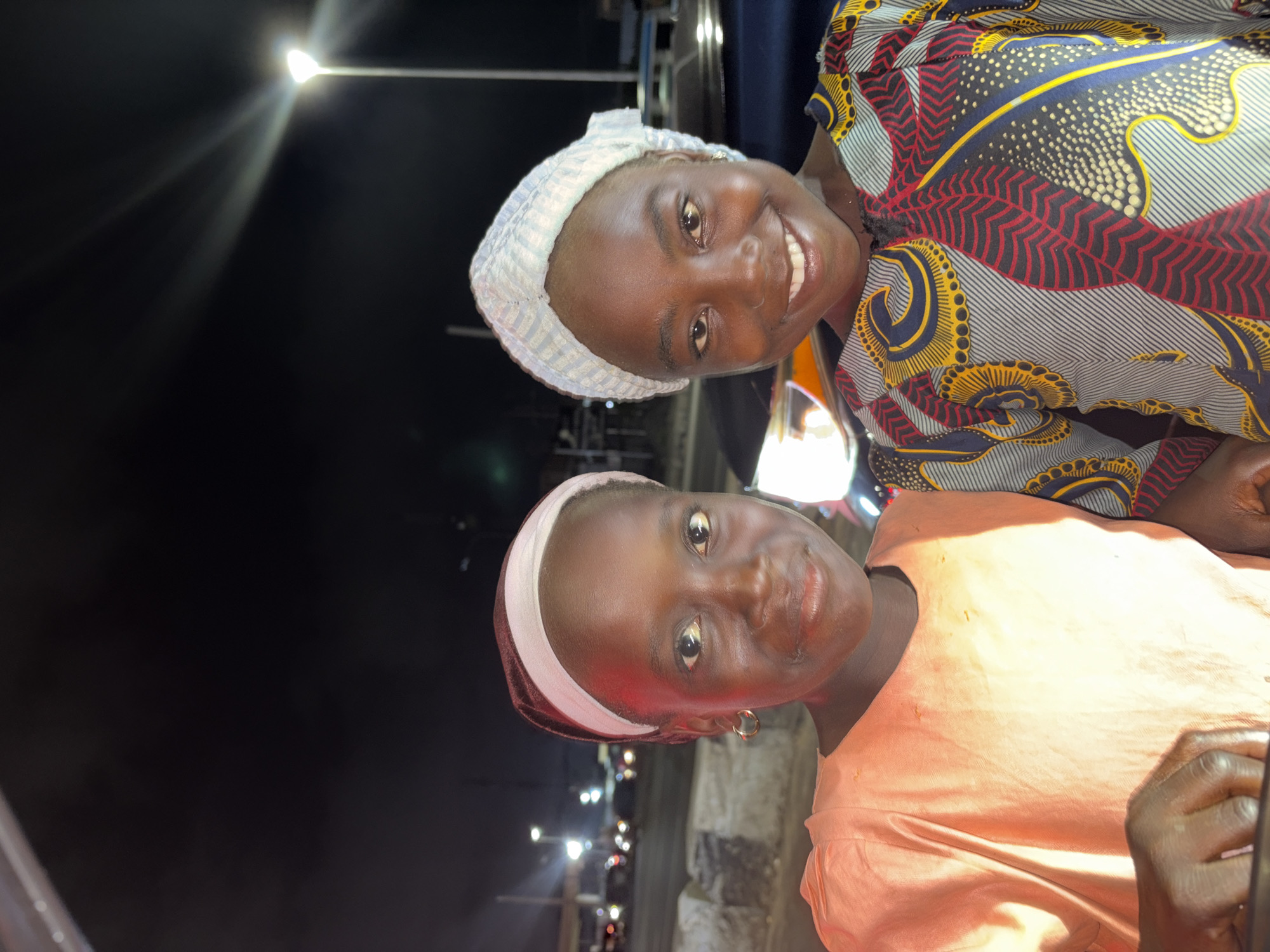 Two dark-skinned women pose for a photo outdoors at night. The woman on top wears a light-colored patterned headscarf and a colorful printed top. The woman below has a dark-colored headscarf and a peach-colored shirt. Street lights illuminate the background, indicating they are in an urban area. Given the location of Alaguntan, Nigeria, the setting is likely a street or public space within the city.