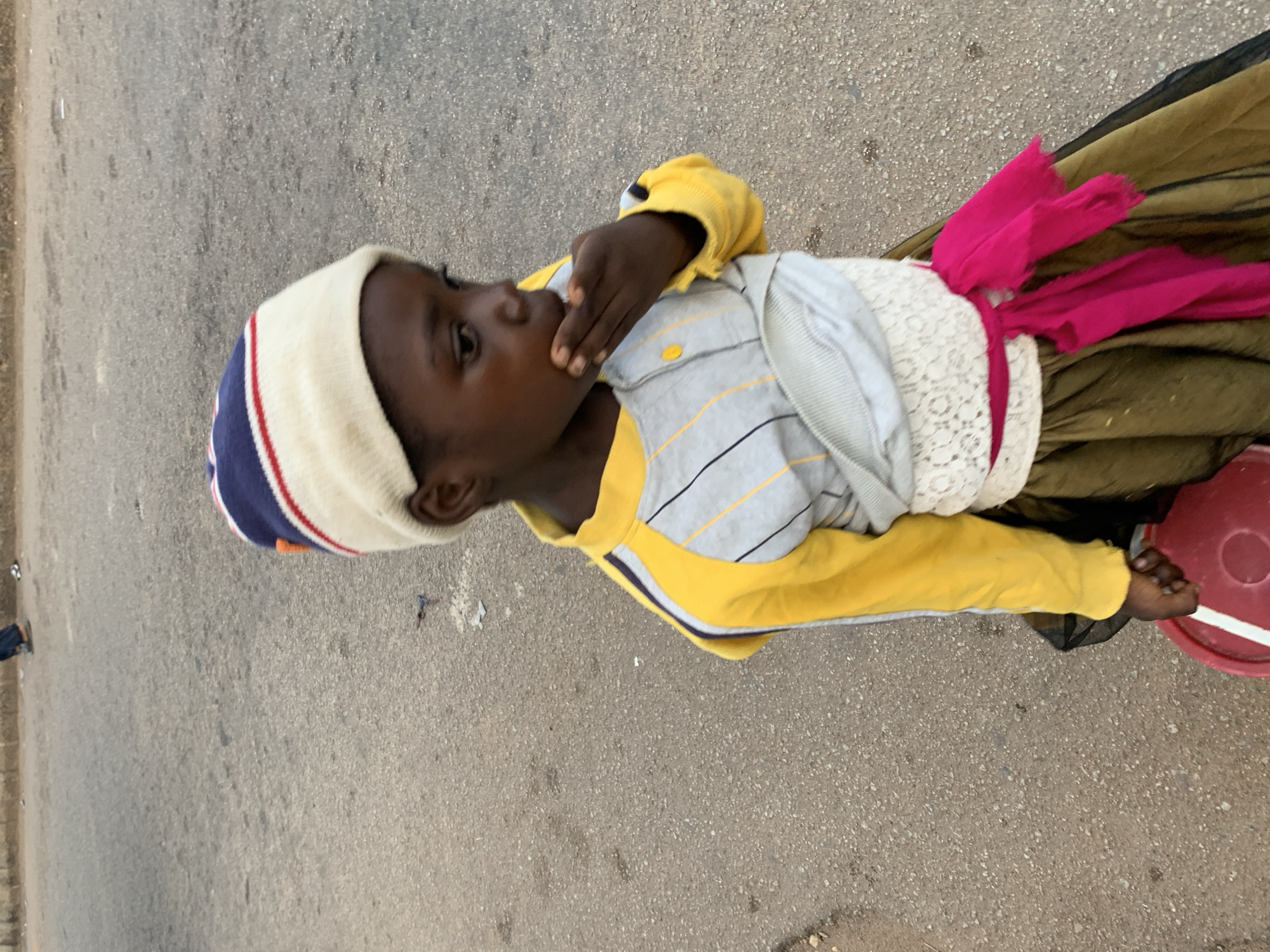 In the city of Jos, Nigeria, a young Black girl stands on an asphalt road. She is wearing a cream-colored knit cap with red and blue stripes. Her clothing includes a yellow and gray long-sleeved shirt with black pinstripes, a white laced sash around her waist, and a dark green and black skirt. A bright pink sash is tied around her waist over the white lace. She is holding a red bucket in her left hand. The girl has her right hand raised to her mouth, and her gaze is directed downwards and to the right. The background is the textured surface of the road, with some debris and faint footprints visible. In the distance, out of focus, two figures can be seen walking on the road.