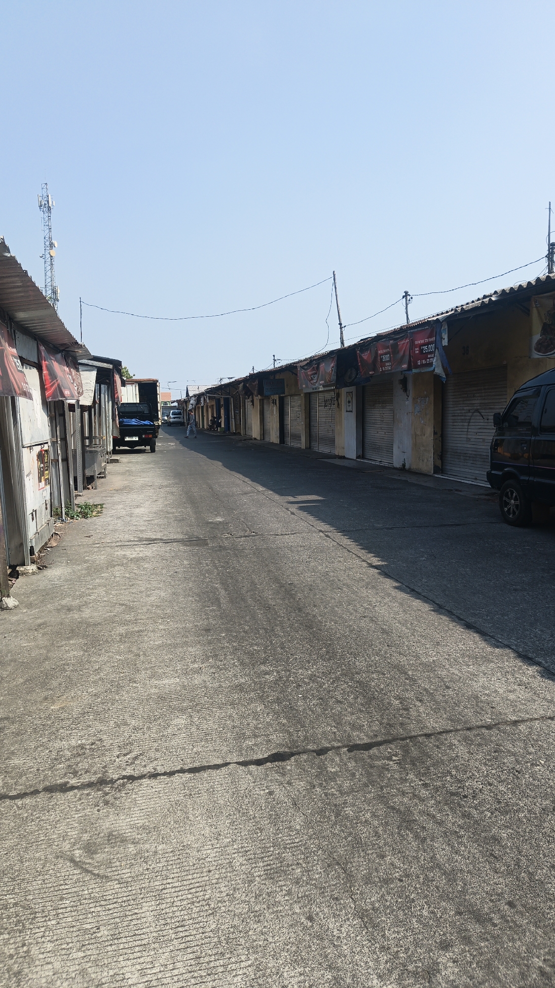This image depicts a street scene in Jekulo, Indonesia. The street is paved with concrete and lined with a row of small, closed shops on the right side of the frame. The shops have metal roller shutters, some of which display advertisements. The buildings appear to be made of wood and corrugated metal, with some showing signs of age and weathering. On the left side of the street, there are more structures, also appearing to be shops or stalls, some of which are open and appear to be in use. A dark-colored van is parked on the right side of the road, and a truck is visible further down the street on the left. A person is walking in the distance on the left side of the street. The sky is clear and blue, indicating a sunny day. A cell tower is visible in the background on the left. The overall impression is of a commercial area, possibly a market or a row of small businesses, during daylight hours.