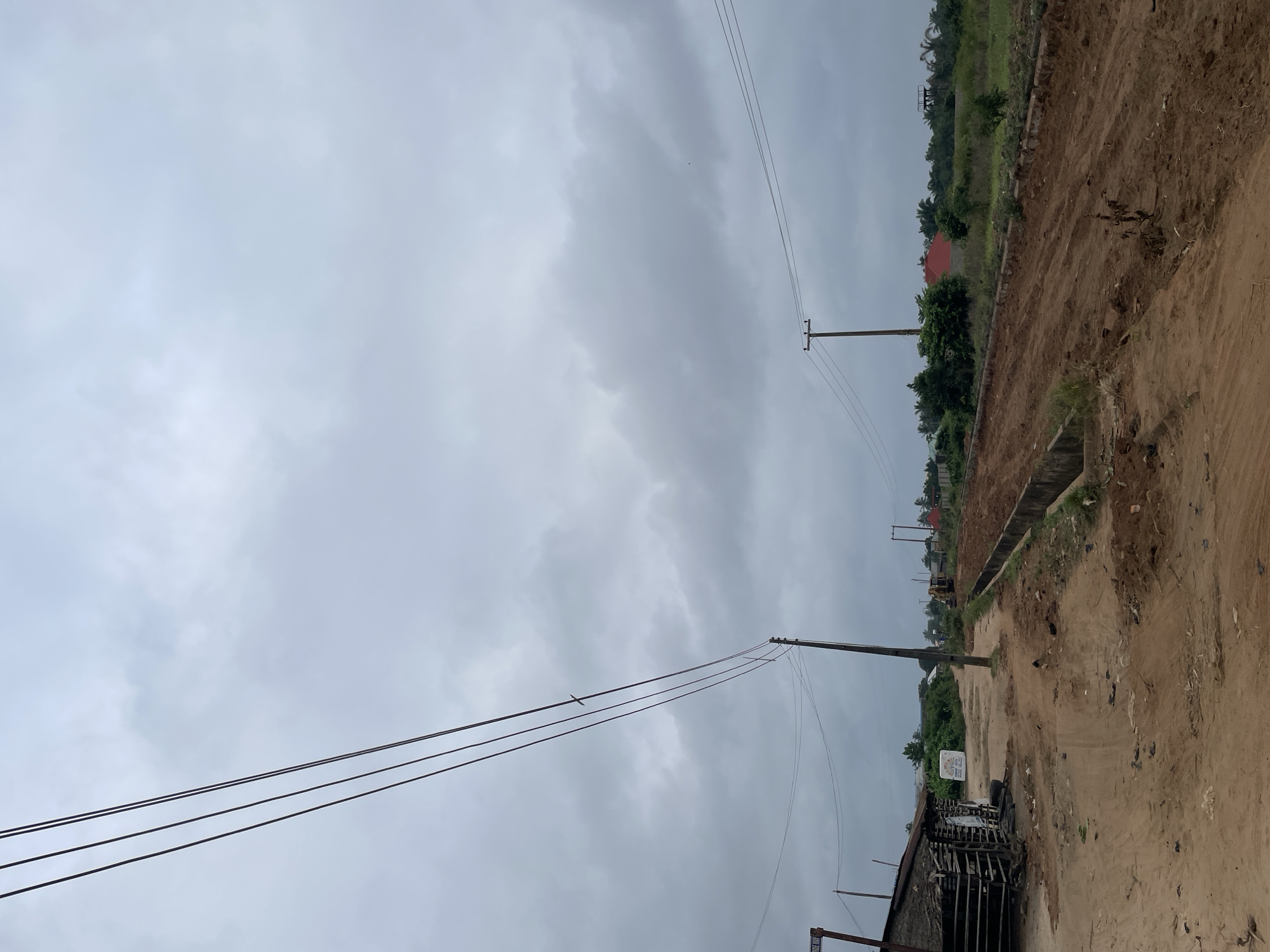 The image depicts a scene in Iyafin, Nigeria, on a cloudy day. A dirt road runs through the center of the frame, with construction or development occurring on the right side, evident from the excavated earth and concrete drainage channels. Power lines are strung across the sky, supported by wooden poles. On the left side of the road, a rudimentary structure with a thatched roof and wooden walls is visible, next to stacked tires and debris. Further down the road, a yellow construction vehicle and various buildings with red roofs can be seen in the distance. Lush green vegetation borders the development area.