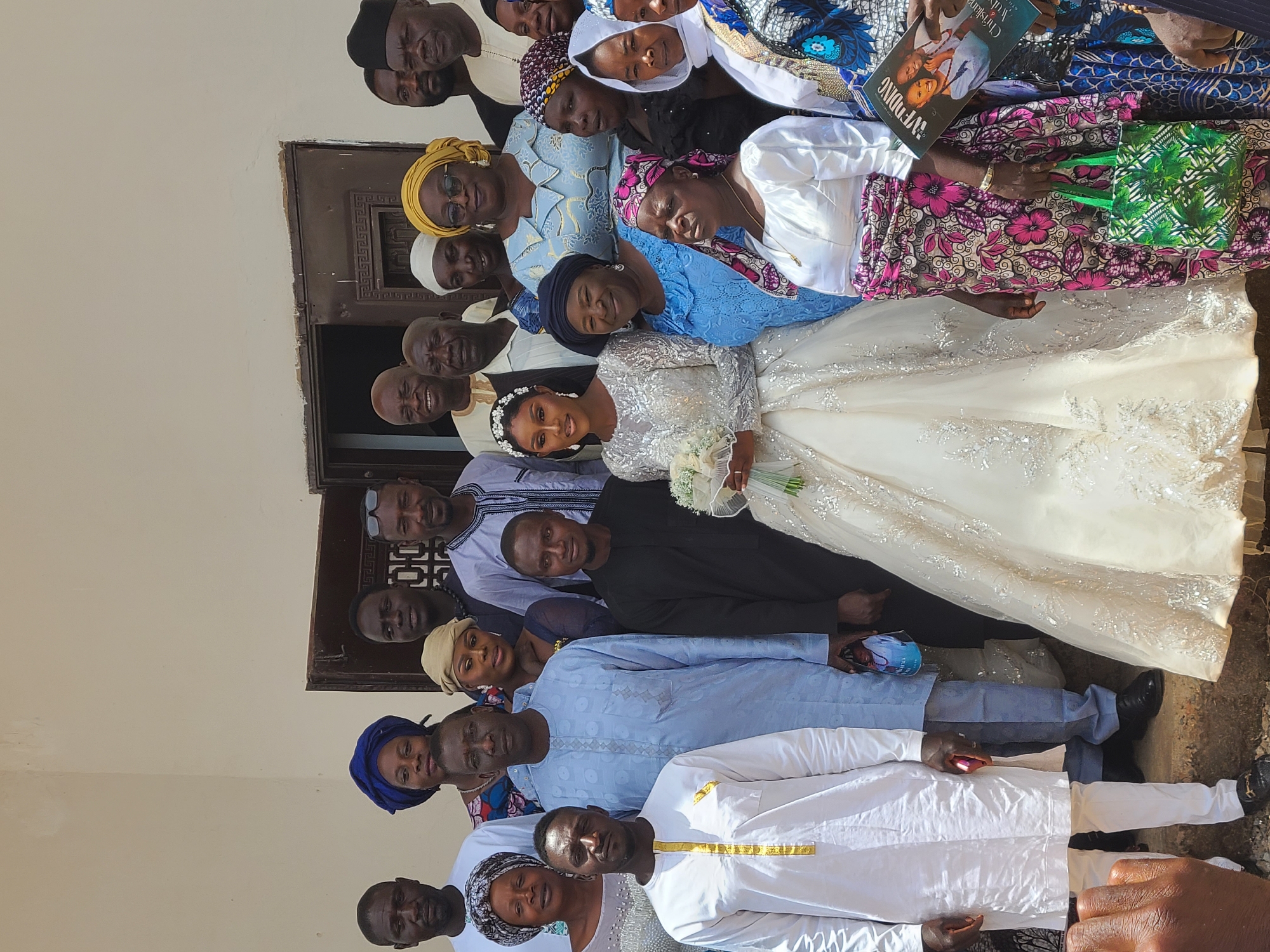 A wedding group photo is captured in Barakin Lerawi, Nigeria. The group consists of about 20 people, including a bride in a white wedding gown and a groom in a black suit. The individuals are standing close together, smiling for the camera. Most of the people are wearing traditional African clothing, with bright colors and patterns. In the background, there's a building with a textured wall. The photo seems to be taken outdoors, possibly near a home. One person in the group is holding a wedding booklet.