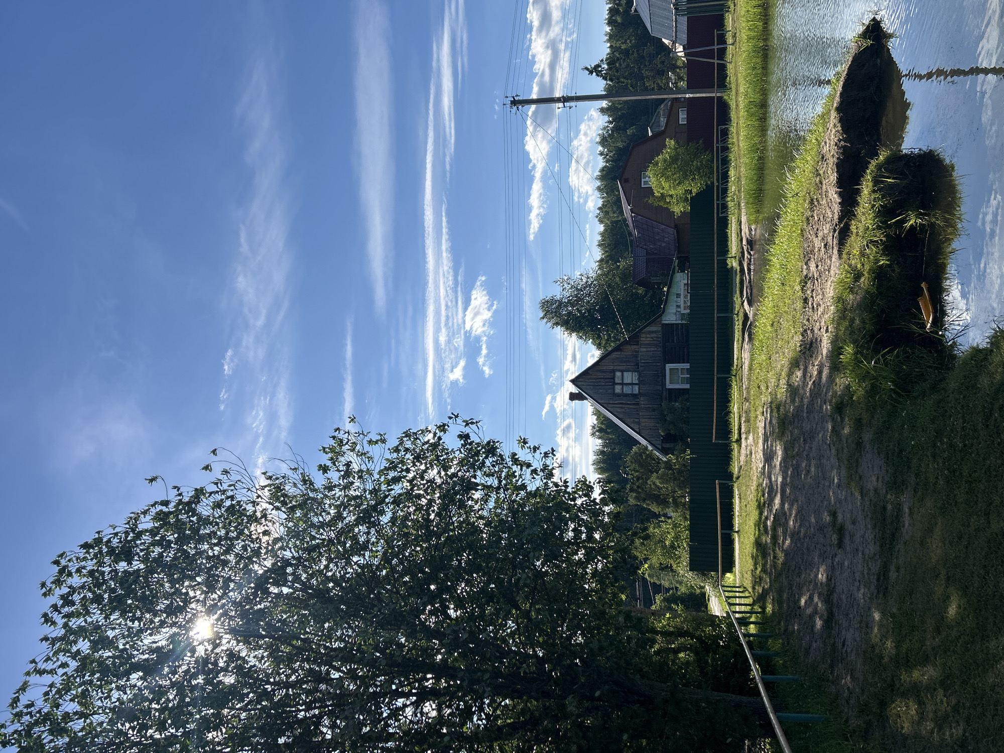 The image depicts a rural scene in the city of Слава Снт, Russia, on a clear day with a bright blue sky adorned with wispy white clouds. A large, leafy tree dominates the left side of the frame, its branches dappled with sunlight. In the background, several wooden houses with peaked roofs are visible, surrounded by dense green foliage and a dark green fence. Power lines stretch across the sky, supported by a utility pole on the right. In the foreground, a body of water reflects the sky, with a sandy bank covered in green grass leading to it. A metal railing with turquoise posts and a wooden top runs along the edge of a dirt path.