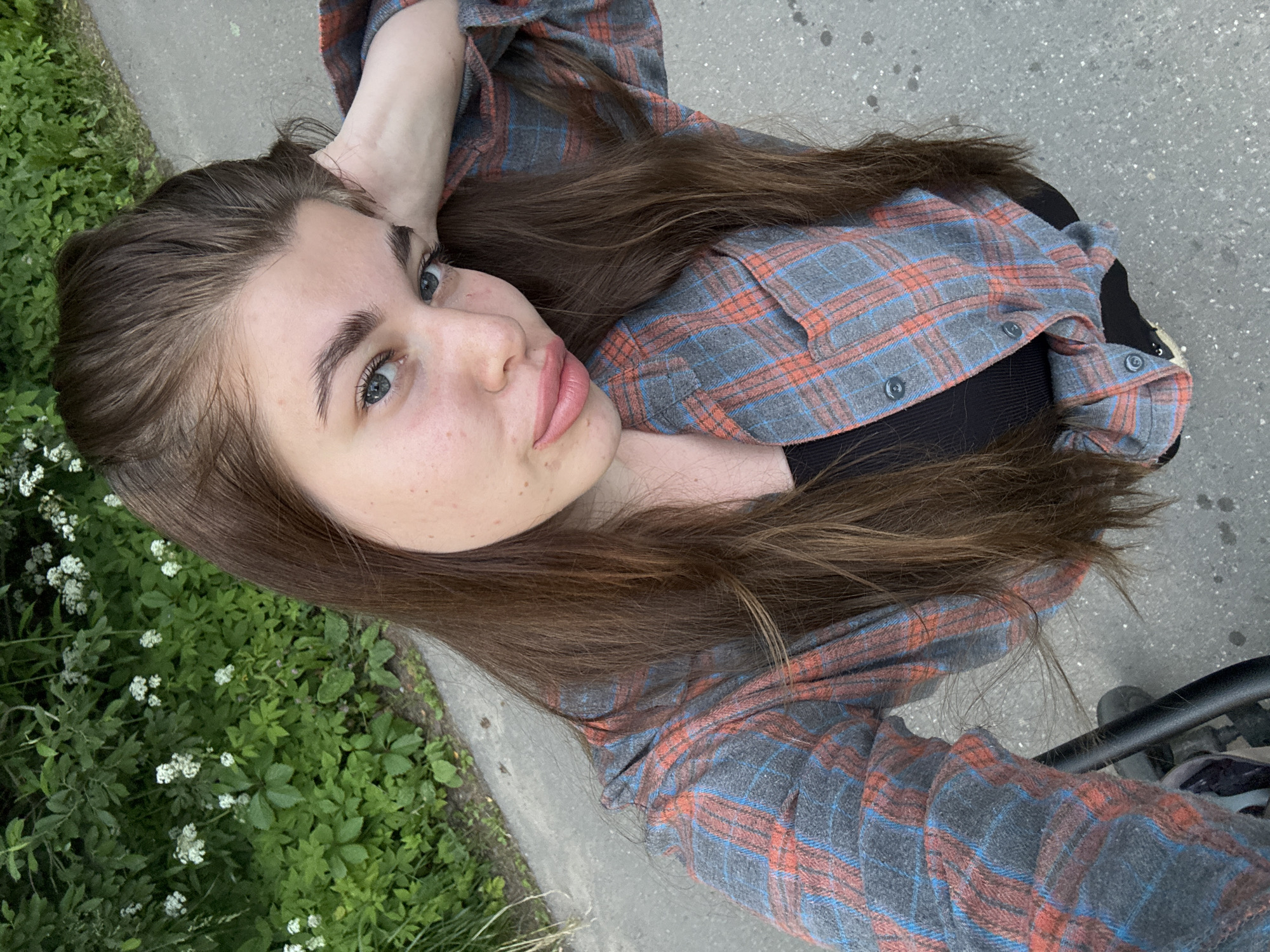 A young woman with long, brown hair is captured in a selfie taken outdoors. She is wearing a grey, red, and blue plaid shirt over a black top. Her left arm is resting on her head. The background consists of lush green foliage with small white flowers, and a grey paved surface, likely a sidewalk or path. The image is focused on the woman's face and upper body. The location is specified as Shevlyagino, Russia.