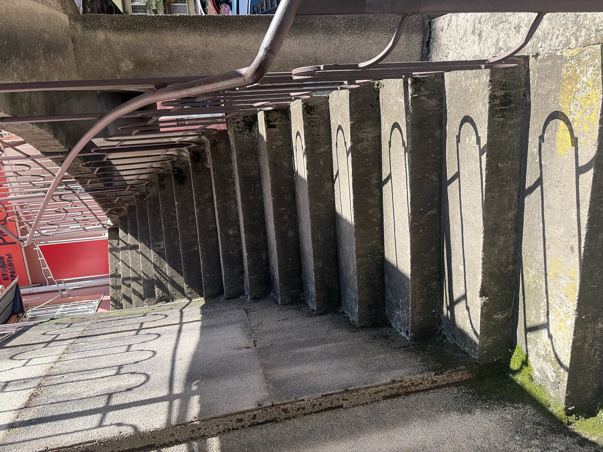 An outdoor concrete staircase is depicted from a high vantage point, looking down along its length. The stairs are constructed from solid, grey concrete, showing signs of age and wear, with patches of green moss or lichen visible on the edges and risers, especially on the right side. A dark metal railing with a distinctive wavy, decorative pattern runs along the left side of the staircase, casting long, intricate shadows onto the steps and the landing below.
The strong, crisp shadows indicate bright, sunny weather, likely during the morning or late afternoon given the angle of the light. The sun highlights the texture of the concrete and the patterns created by the railing's shadows.
In the background, to the left of the stairs, part of a building or structure with a large red facade is visible. White metal framework and railings are also seen in this area. Faint Russian text, including "СТУДИИ КРАСОТЫ" (Beauty Salon) and "РАБОТЫ" (Works), is discernible on the red surface, suggesting a commercial establishment. Further details of urban architecture, such as windows and an air conditioning unit, are partially visible in the upper background. No people are present, and no specific activity is taking place, lending a quiet, observational quality to the scene within an urban setting in Sochi, Russia.